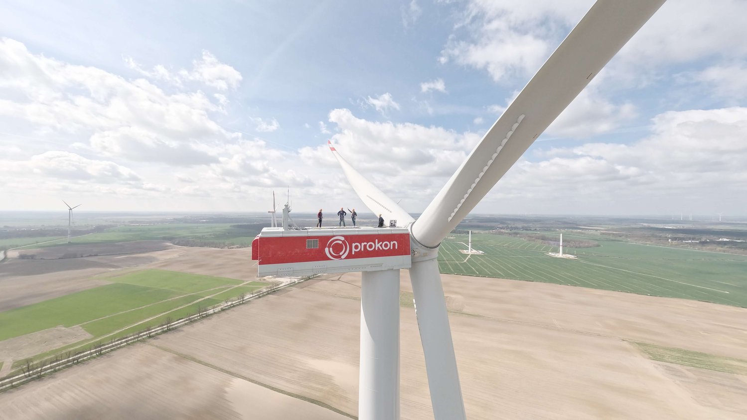 Vier Personen stehen auf einer großen Windturbine mit der Aufschrift „Prokon“ und blicken unter einem teilweise bewölkten Himmel auf Felder und andere Windturbinen.