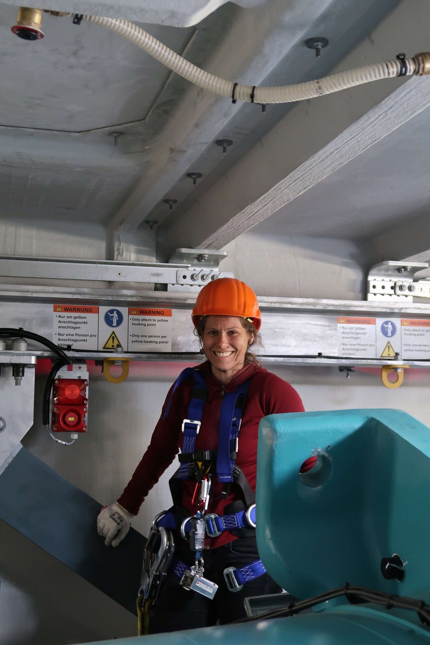 Eine Person mit Sicherheitsgurt, Handschuhen und orangefarbenem Schutzhelm steht im Innenbereich in der Nähe von Industrieanlagen und Sicherheitswarnschildern.