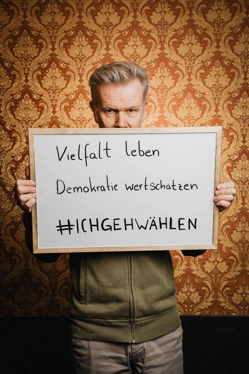 Person hält vor gemustertem Hintergrund ein Schild mit deutschem Text, der für Vielfalt und Demokratie wirbt.