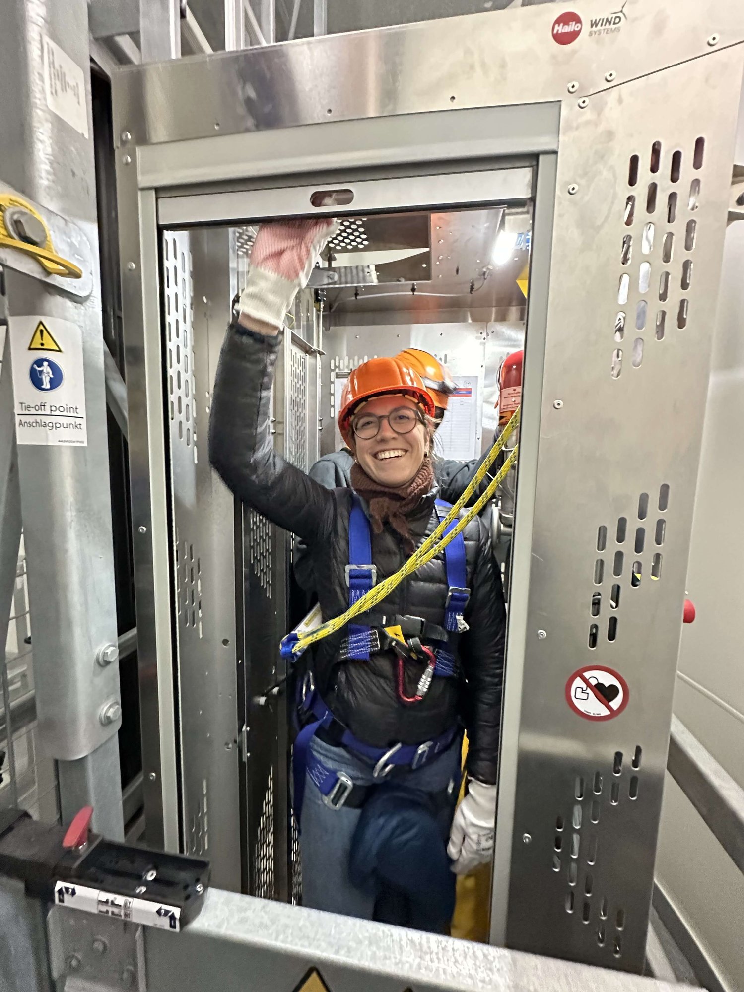 Eine Person in Schutzausrüstung und Helm steht in einem industriellen Metallaufzug oder -gehäuse und lächelt in die Kamera. Um sie herum sind Sicherheitsschilder und -ausrüstung sichtbar.