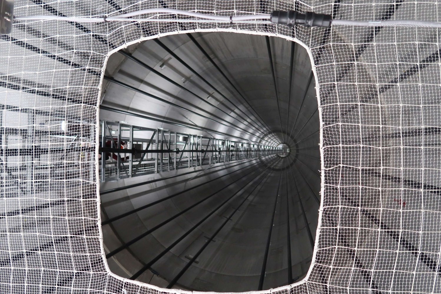 Blick durch ein quadratisches Maschennetz in einen langen, zylindrischen, metallischen Tunnel mit symmetrischen Linien und einem Fluchtpunkt in der Ferne.