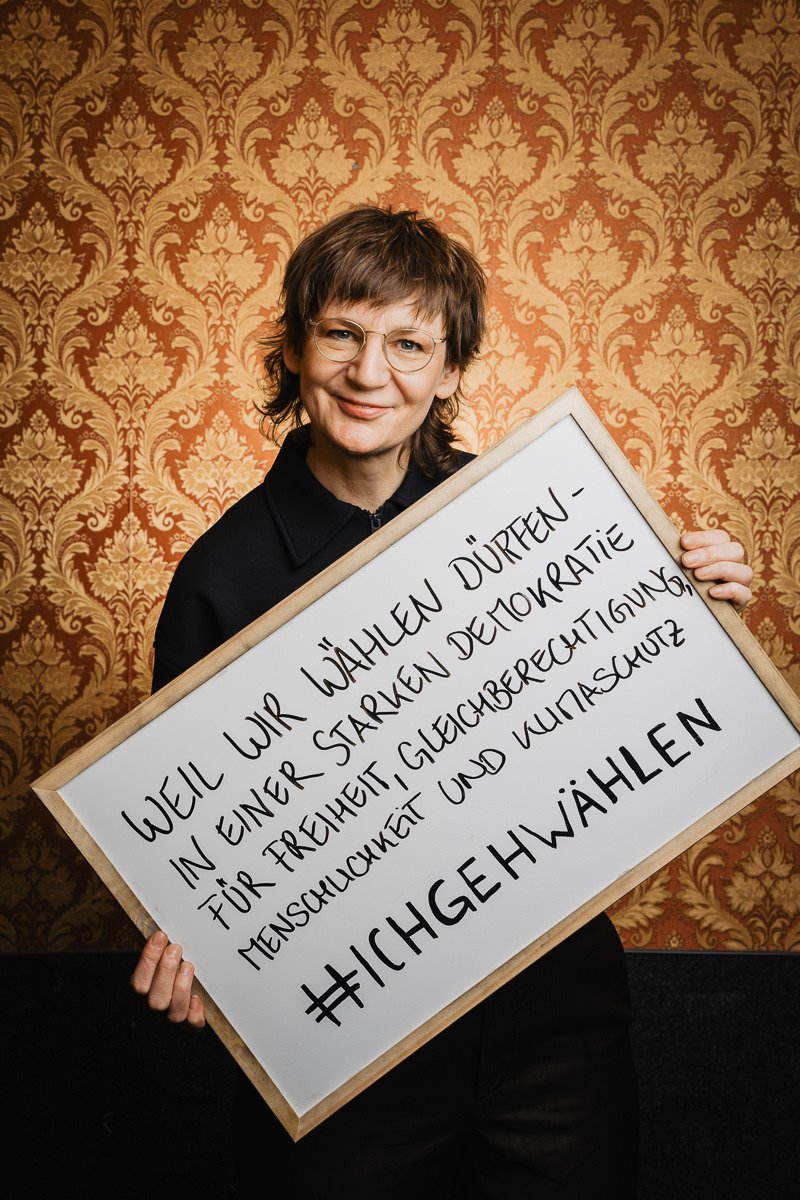 Person hält ein Schild mit handgeschriebenem deutschen Text über Wahlen und die Unterstützung von Gleichheit, Demokratie und Menschenrechten. Der Hintergrund ist eine gemusterte Tapete.