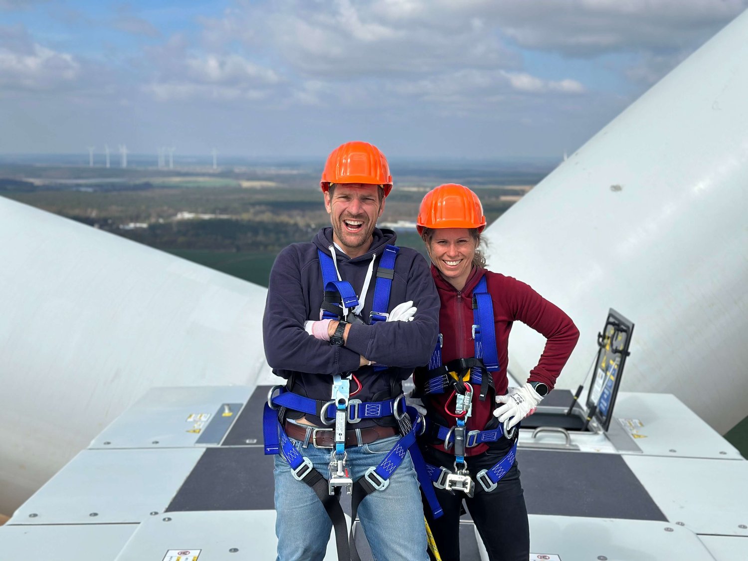Zwei Personen mit Sicherheitsgurten und orangefarbenen Helmen stehen auf einer Windkraftanlage mit großen Rotorblättern und einer Landschaft im Hintergrund.