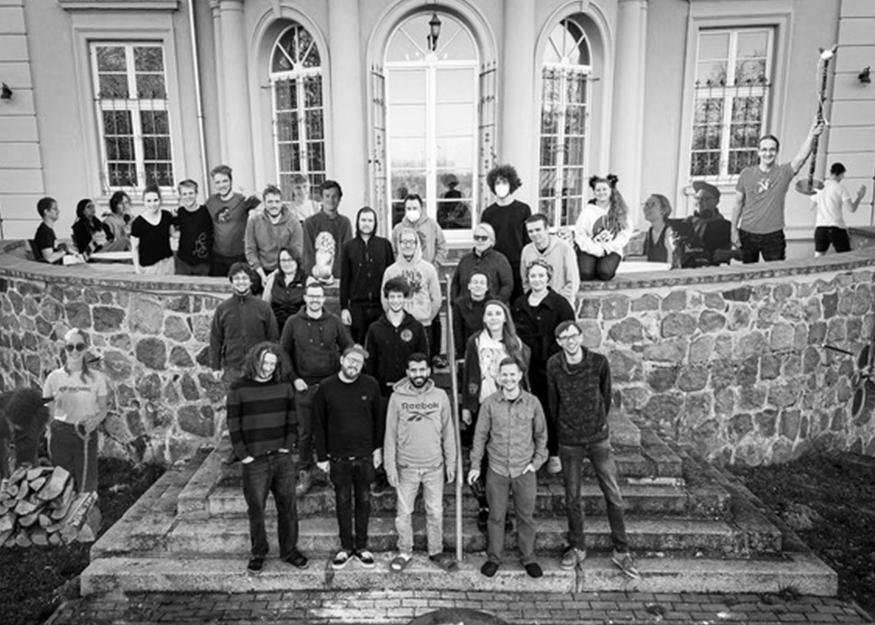 Eine Gruppe von Menschen steht und sitzt auf einer Steintreppe vor einem großen Gebäude mit Bogenfenstern. Das Bild ist schwarz-weiß.