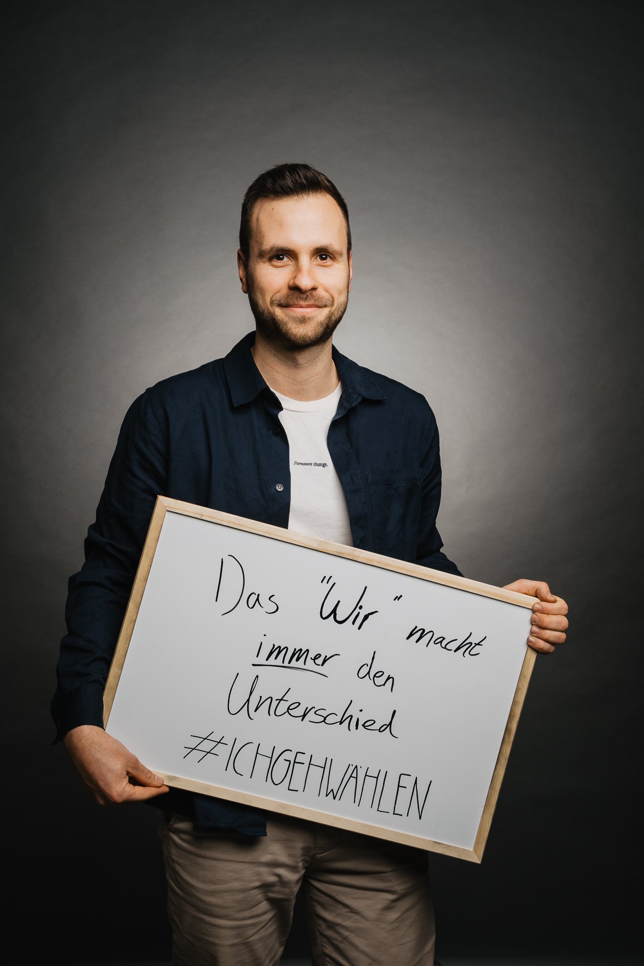 Mann hält ein Whiteboard mit handgeschriebenem Text auf Deutsch: „Das ‚Wir‘ macht immer den Unterschied #ICHGEHWAHLEN“ und steht vor einem dunklen Hintergrund.