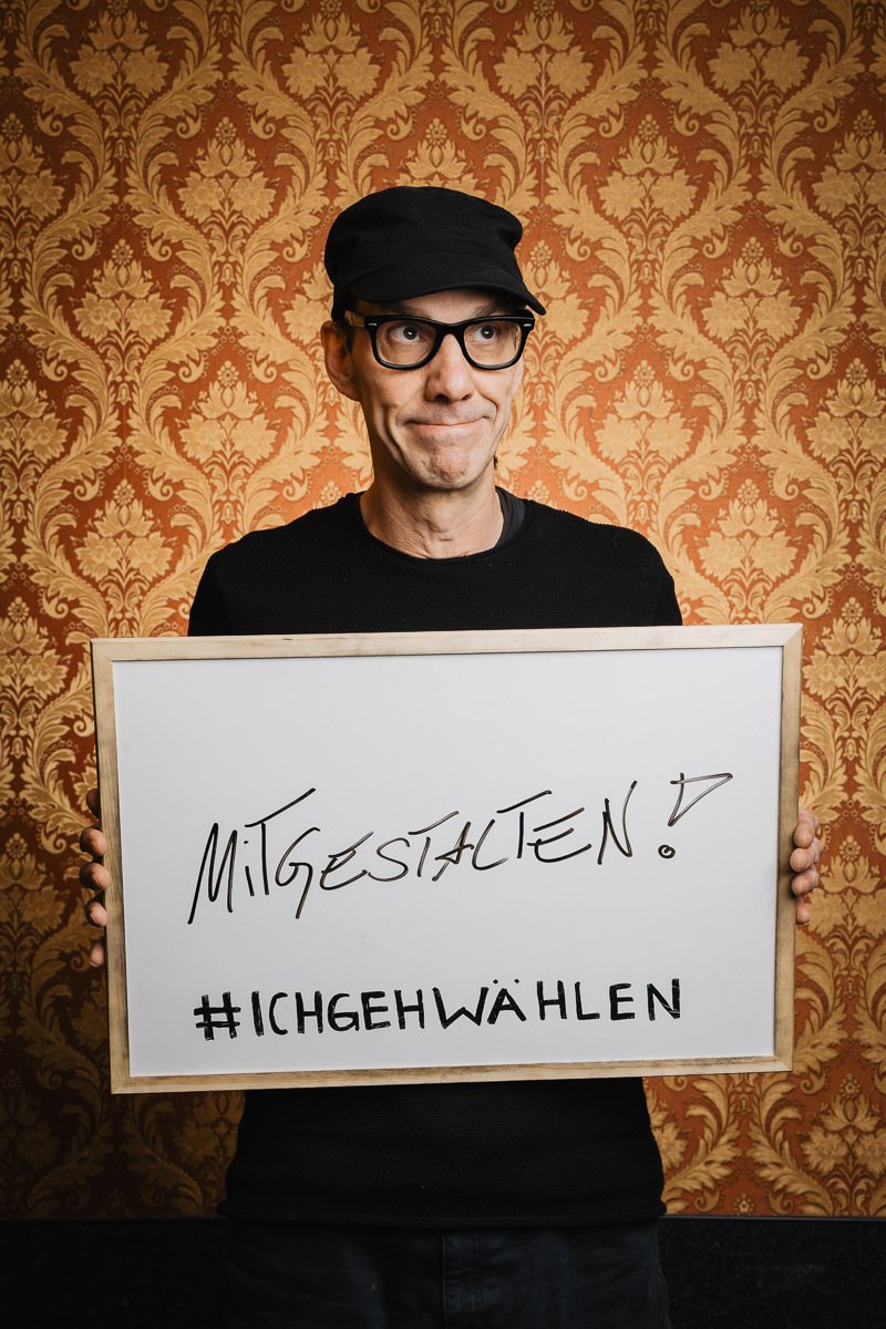 Person mit einem Schild mit der Aufschrift „Mitgestalten? #Ichgehwählen“ vor gemusterter Tapete.