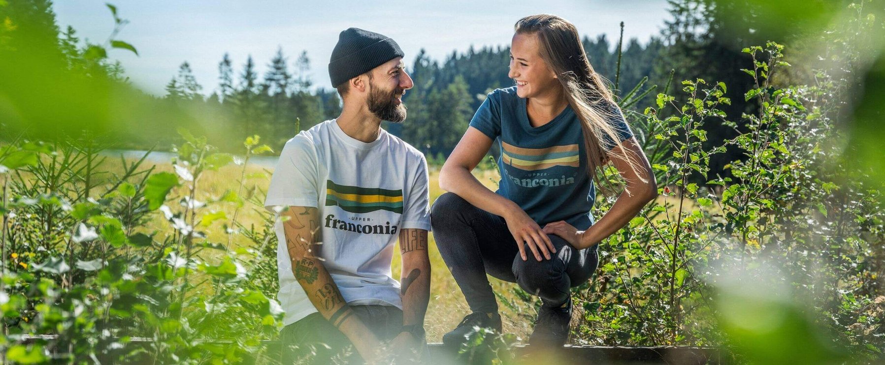 Zwei Personen in „Upper Franconia“-T-Shirts, eine kniend und eine hockend, lächeln sich in einer grünen Außenumgebung mit Bäumen im Hintergrund an.