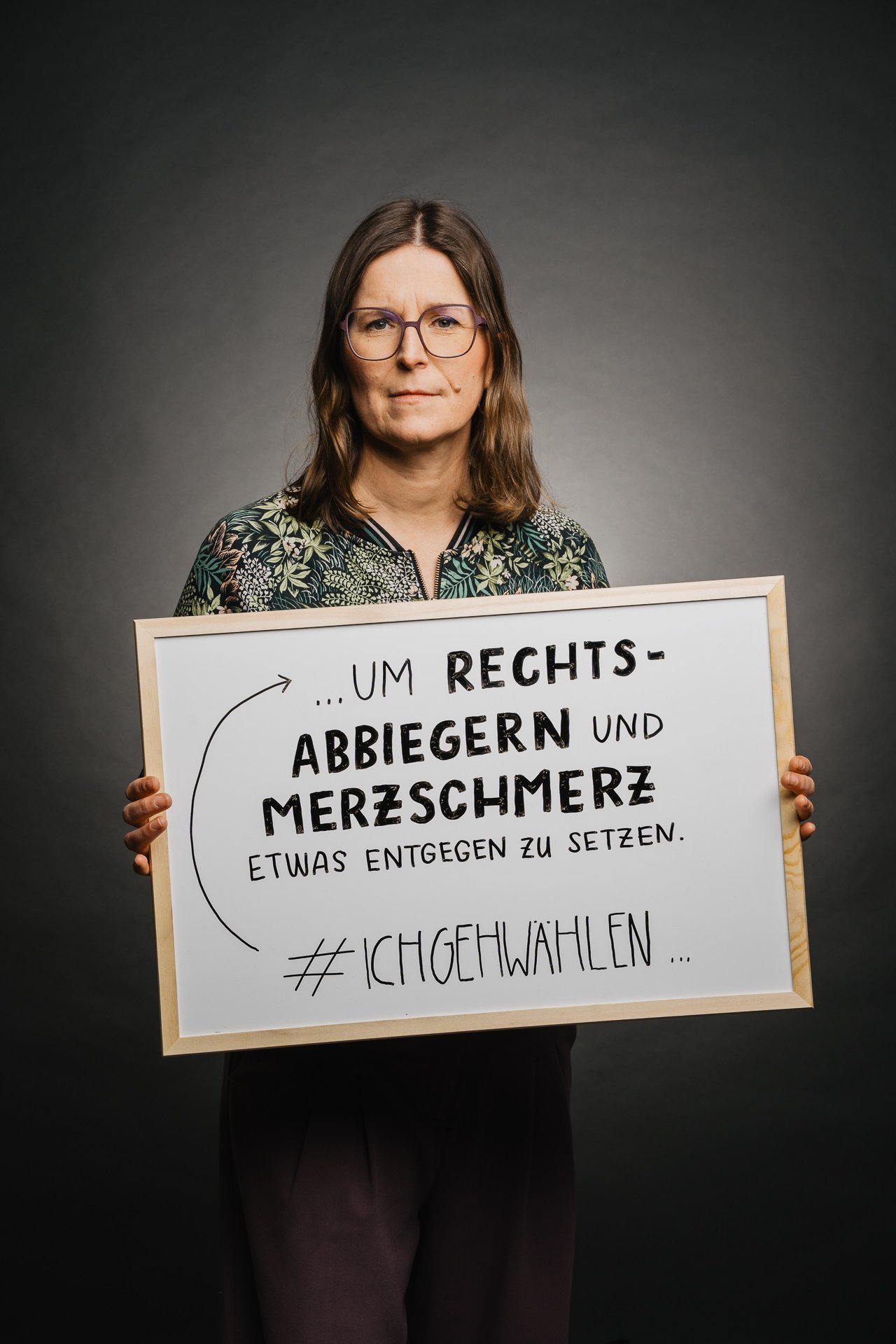 Eine Person hält ein Schild mit deutschen Texten zu sozialen Themen, darunter „Rechts-Abbiegern“ und „Merzschmerz“, gefolgt von „#ICHGEHWAHLEN“. Sie steht vor einem dunklen Hintergrund.