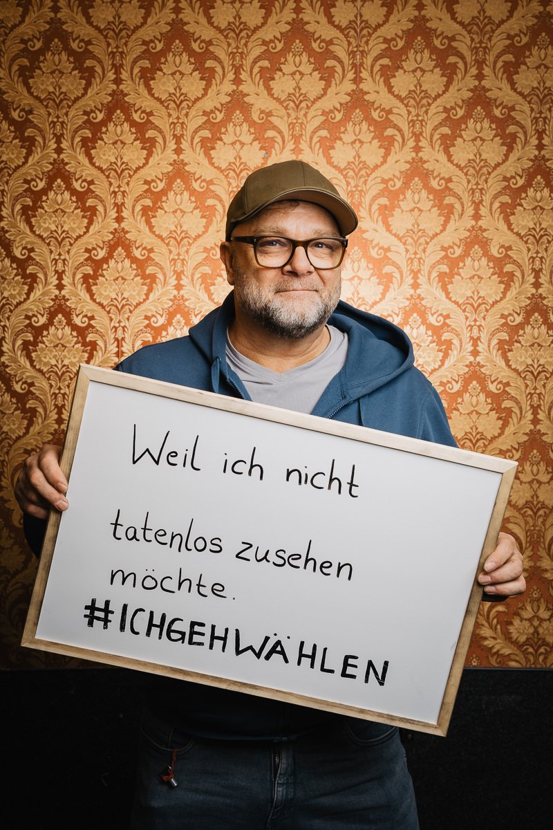 Mann mit Brille und Mütze hält ein Schild auf Deutsch mit der Aufschrift „Weil ich nicht tatenlos zusehen möchte. #ICHGEHWÄHLEN“ vor einem gemusterten Tapetenhintergrund.