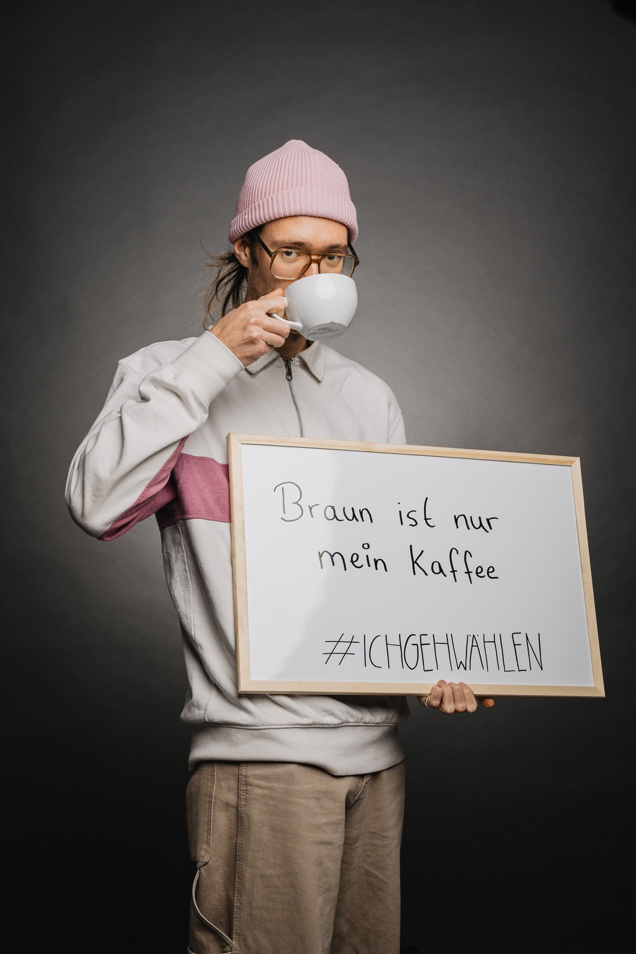 Person mit rosa Mütze und Brille hält ein Schild mit der Aufschrift „Braun ist nur mein Kaffee #ICHEHWAHLEN“ in der Hand, während sie aus einer Tasse trinkt.