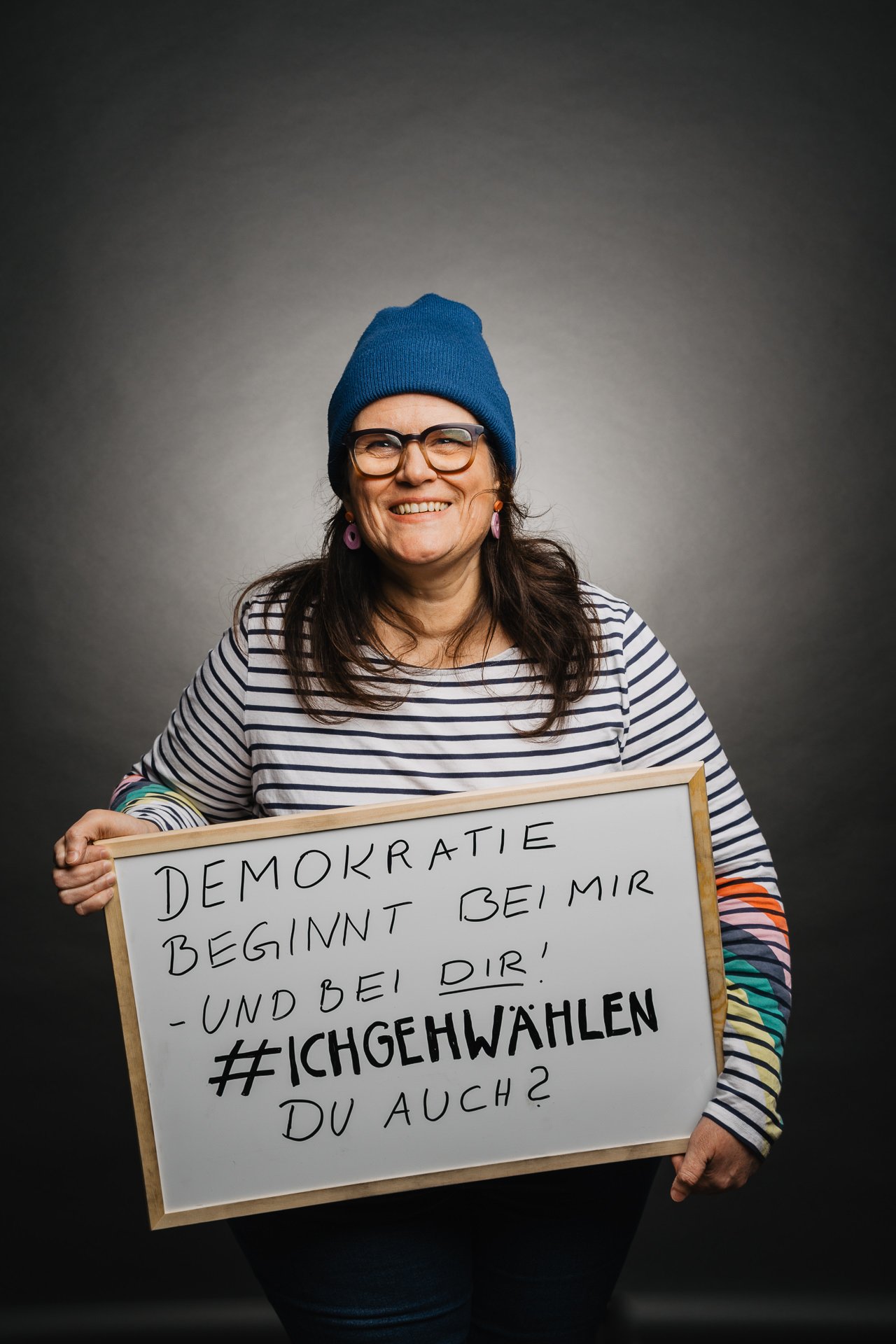 Eine Person mit blauer Mütze und gestreiftem Hemd hält ein Schild mit einem deutschen Text über Demokratie und Wahlen.