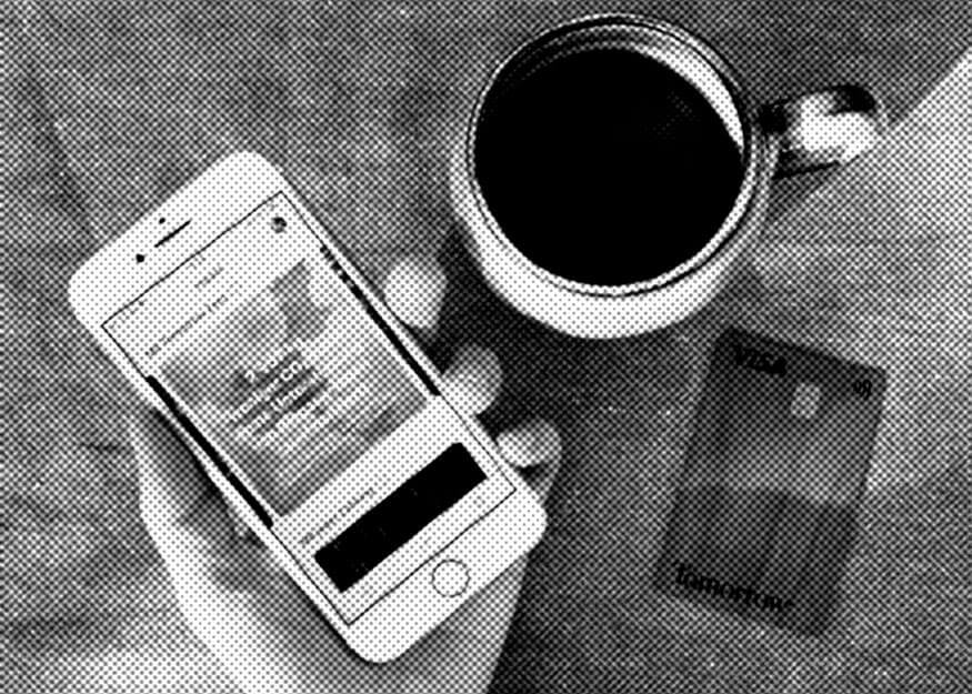 Eine Hand hält ein Smartphone. Auf einem Tisch liegen eine Metalltasse Kaffee und eine Tomorrow Bankkarte.