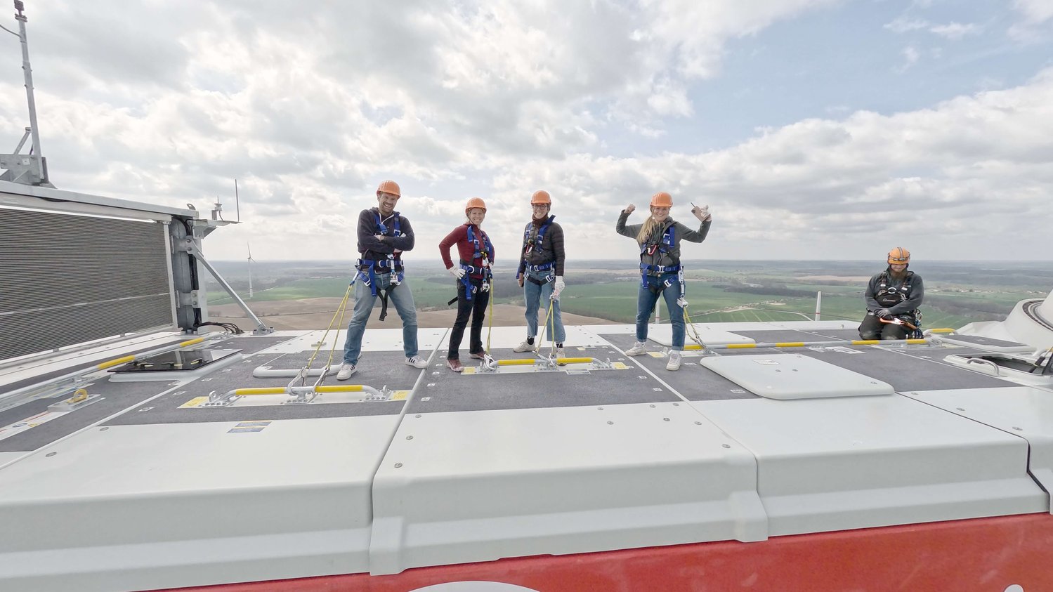 Vier Personen mit Gurten und Helmen stehen auf einer Dachplattform und posieren für ein Foto, während eine fünfte Person allein auf der rechten Seite sitzt. Im Hintergrund sind Himmel und Landschaft zu sehen.