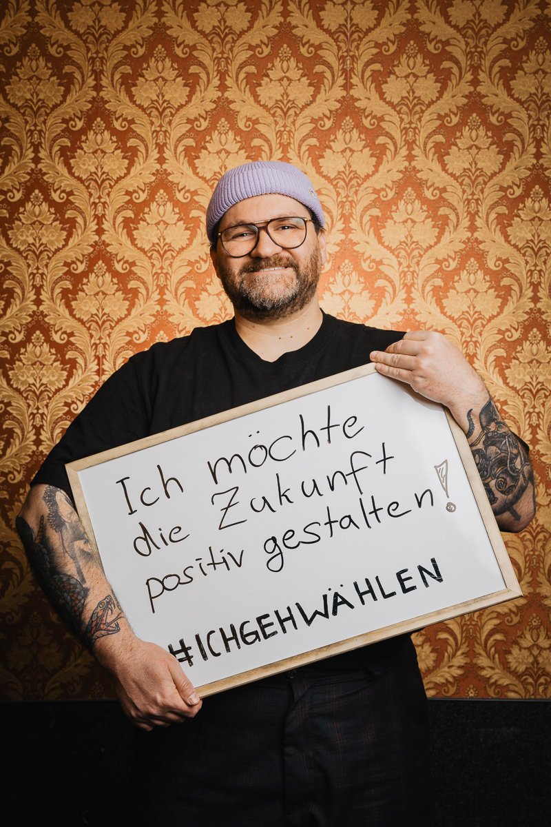 Eine Person mit lila Mütze und Brille hält vor einem kunstvoll gemusterten Hintergrund ein Schild mit der Aufschrift „Ich möchte die Zukunft positiv gestalten! #ICHGEHWÄHLEN“ in der Hand.