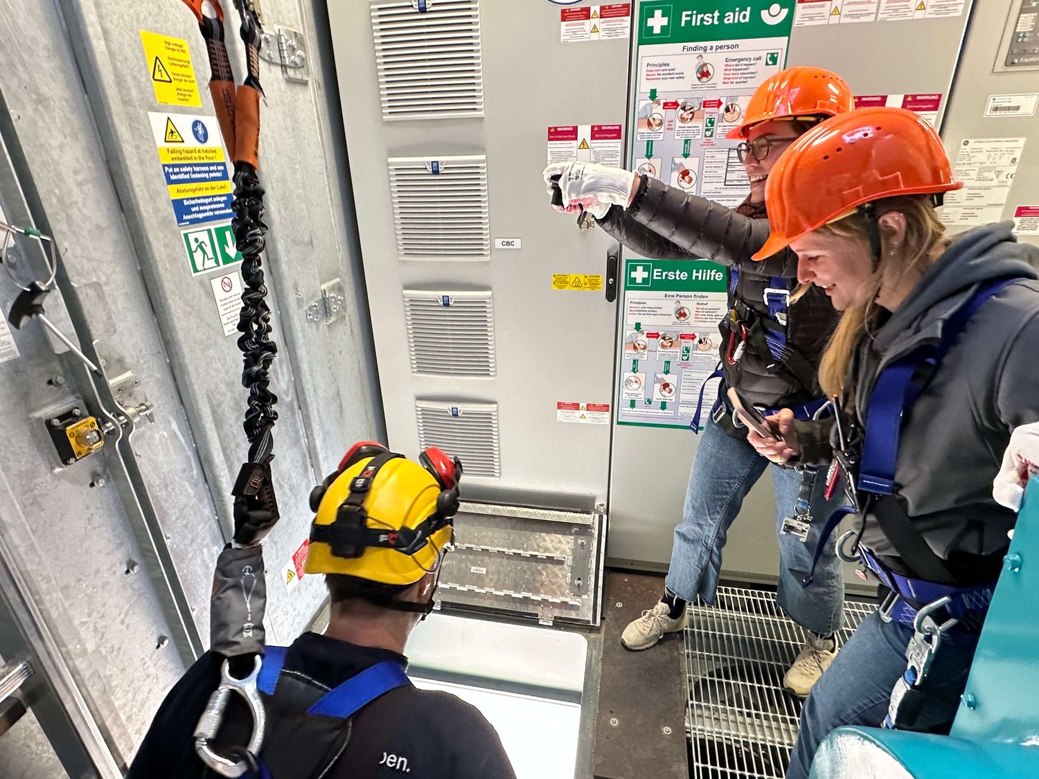 Drei Personen in Schutzausrüstung und Helmen überprüfen die Ausrüstung in einer Industrieanlage in der Nähe von Erste-Hilfe-Schildern und Sicherheitsanweisungen.