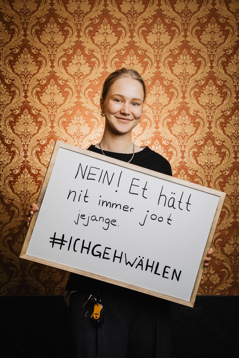 Person hält ein Schild mit dem deutschen Text „NEIN! Et hätt nit immer joot jejange. #ICHGEHWÄHLEN“ vor einem gemusterten Hintergrund.