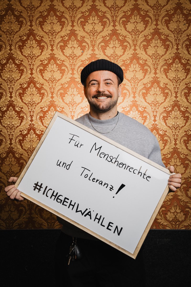 Mann hält ein Schild mit der Aufschrift „Für Menschenrechte und Toleranz! #ICHGEHWÄHLEN“ vor einem kunstvoll gemusterten Hintergrund.