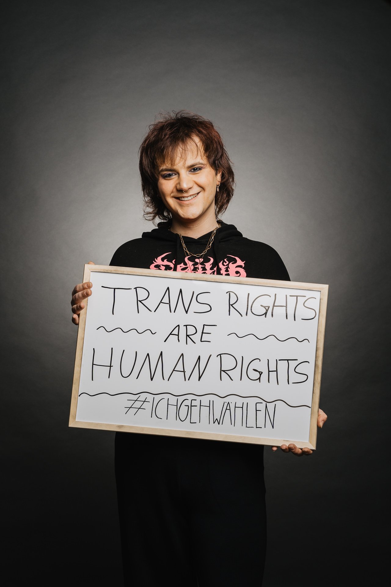 Person, die vor dunklem Hintergrund ein Schild mit der Aufschrift „Trans-Rechte sind Menschenrechte #IchGeheWählen“ hält.