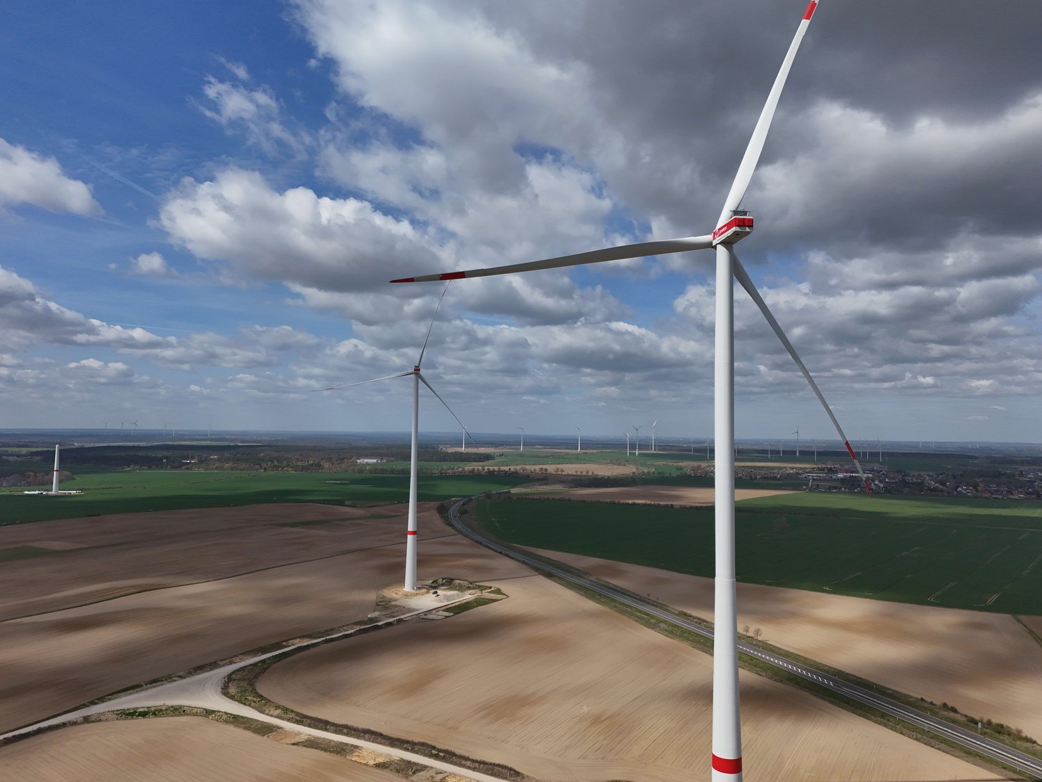 Zwei große Windturbinen stehen auf offenen landwirtschaftlichen Feldern unter einem teilweise bewölkten Himmel. In der Ferne sind weitere Turbinen zu sehen.