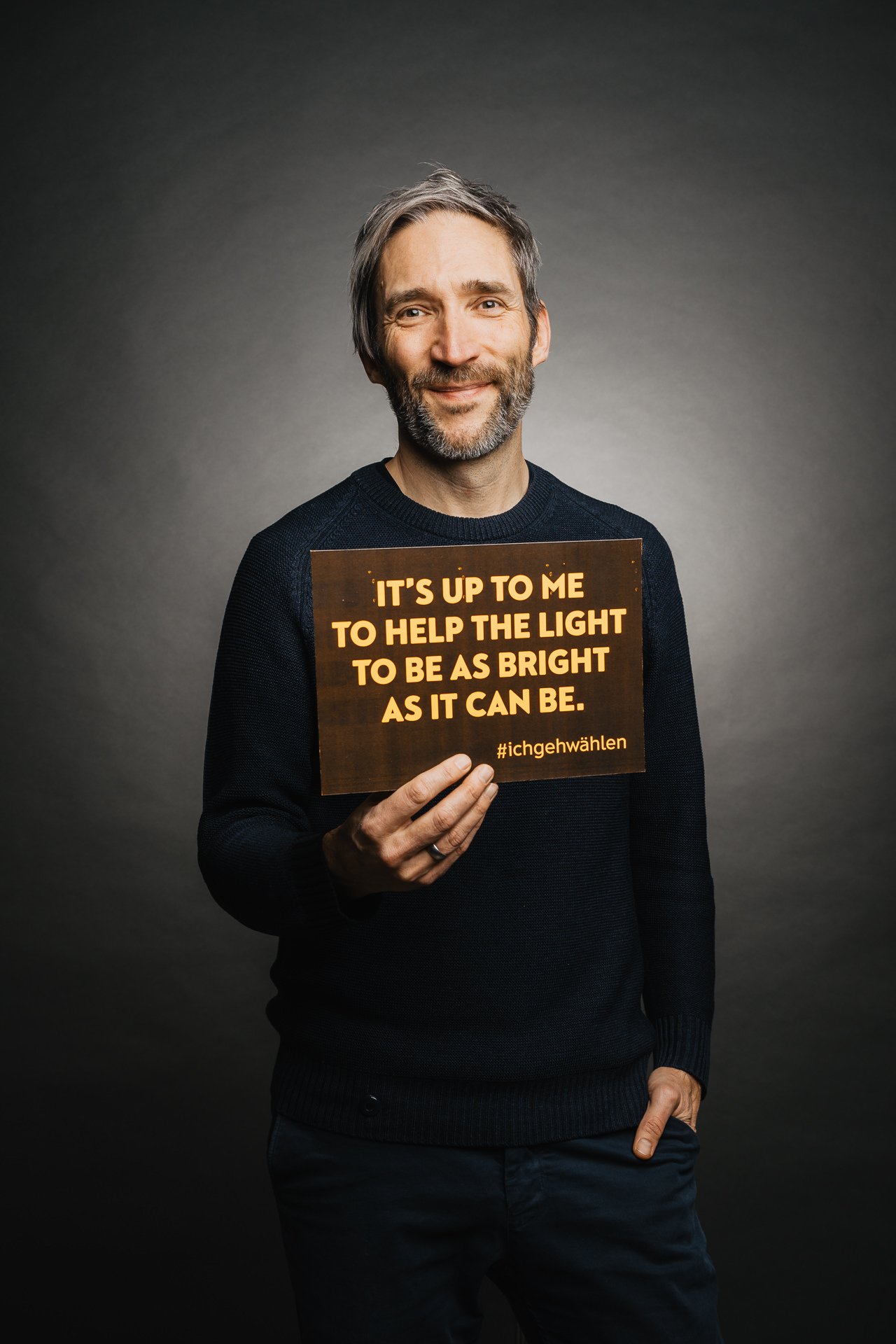 Ein Mann mit Bart und dunklem Pullover hält ein Schild mit der Aufschrift „Es liegt an mir, dafür zu sorgen, dass das Licht so hell wie möglich ist.“.