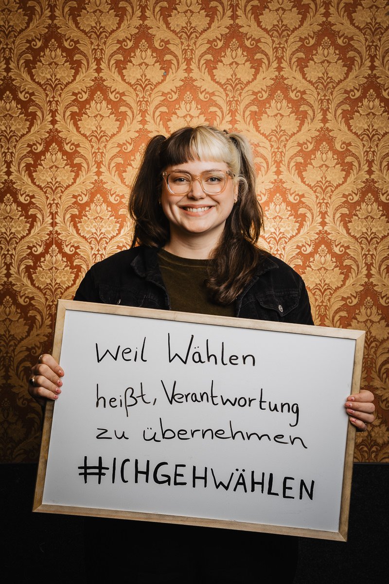 Lächelnde Person mit Brille, die ein Schild mit deutscher Aufschrift hochhält, das zum Wählen aufruft. Im Hintergrund ist eine gemusterte Tapete zu sehen.