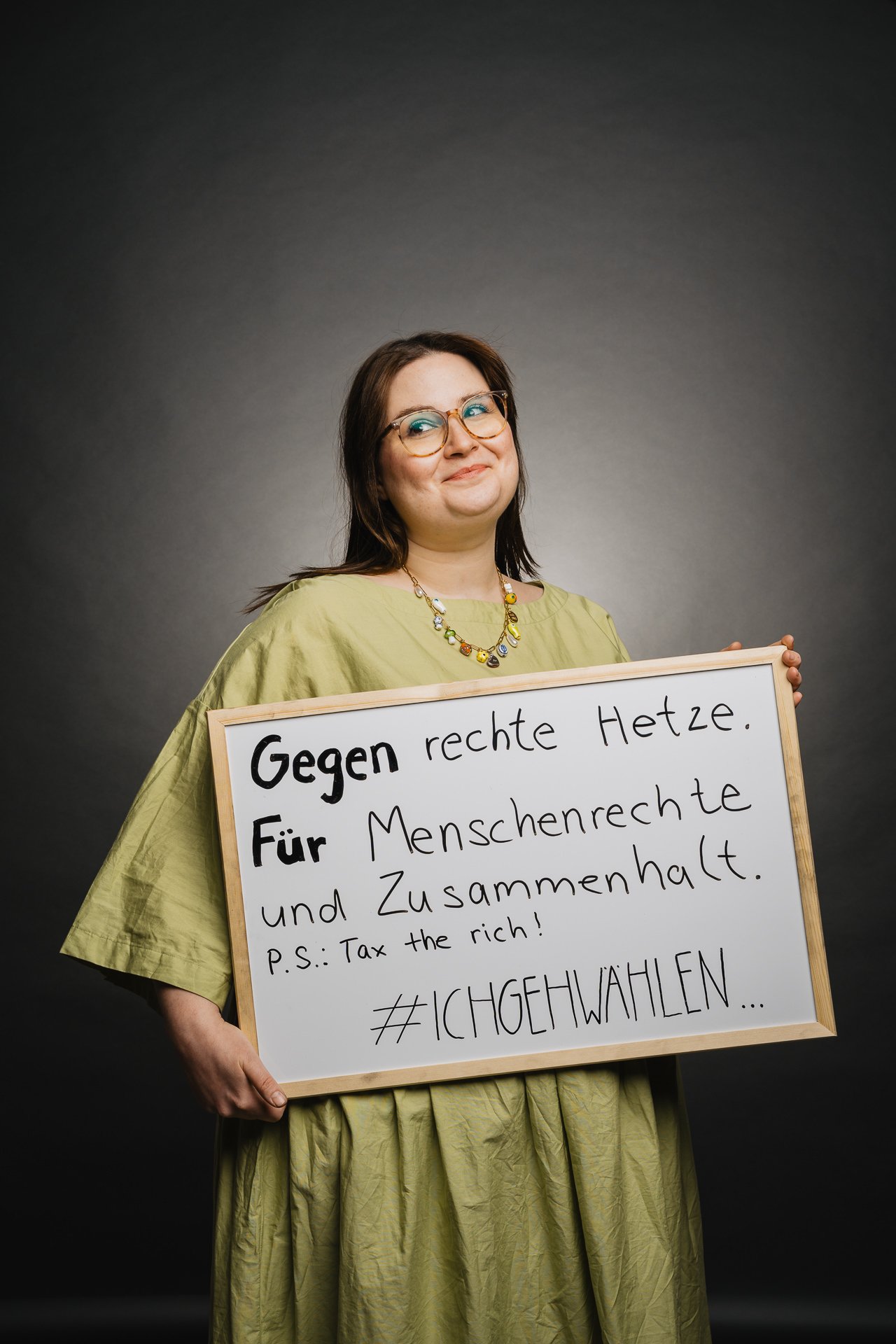 Eine Person in einem grünen Kleid hält vor dunklem Hintergrund ein Schild mit der Aufschrift „Gegen rechte Hetze. Für Menschenrechte und Zusammenhalt. P.S. Tax the rich! #ICHGEHWAHLEN“.