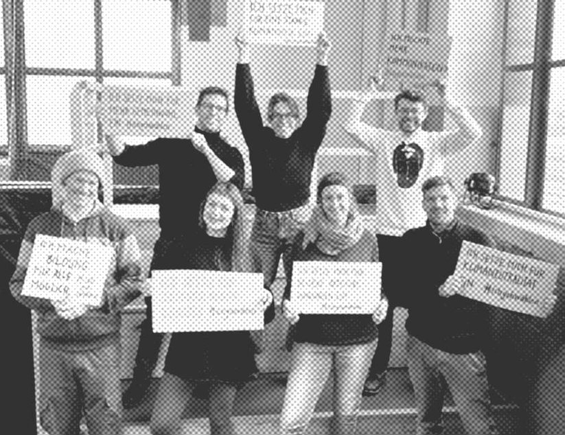 Eine Gruppe von acht Personen posiert im Innenbereich und halten Schilder mit Text.