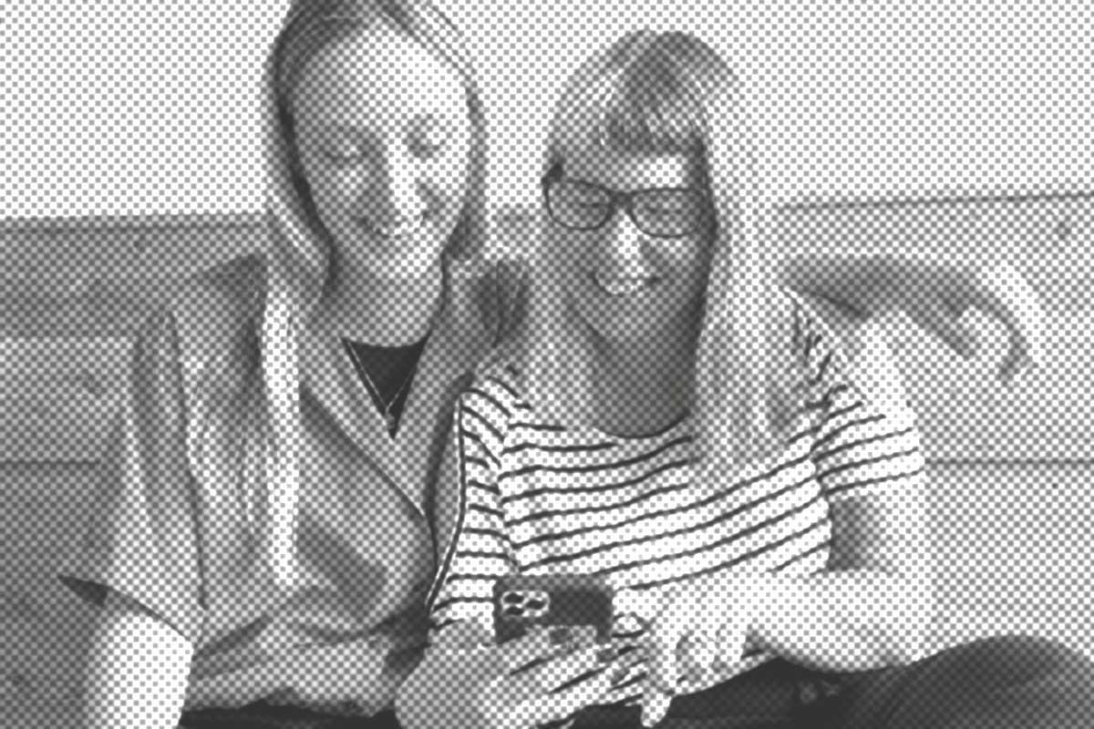 Zwei Personen sitzen lächelnd zusammen und schauen auf ein Smartphone. Eine hat lange Haare, die andere trägt eine Brille und ein gestreiftes Hemd.