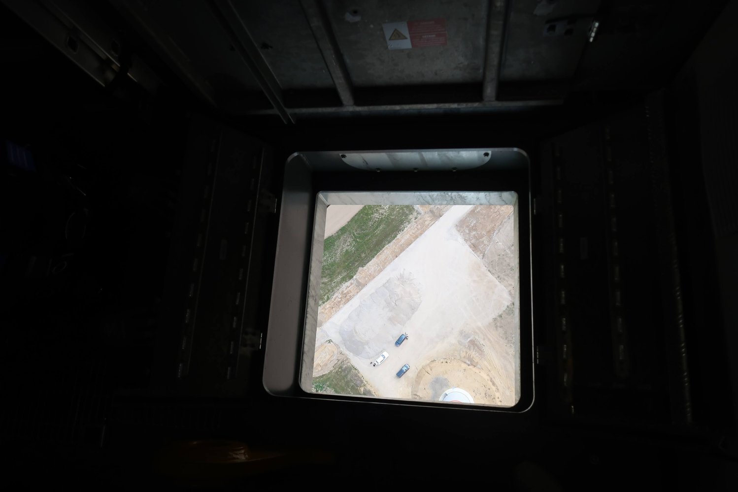 Blick von oben durch ein quadratisches Fenster auf eine Baustelle mit Fahrzeugen auf einem Erd- und Kiesplatz.