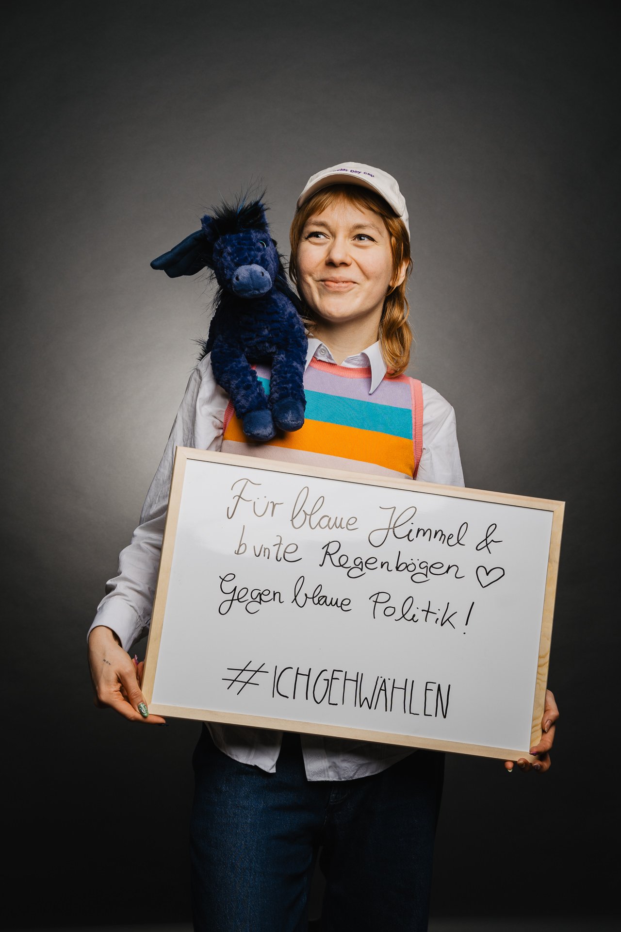 Person, die vor einem dunklen Hintergrund ein Schild mit einem ausgestopften blauen Esel auf der Schulter hält, eine bunte Weste und Mütze trägt.
