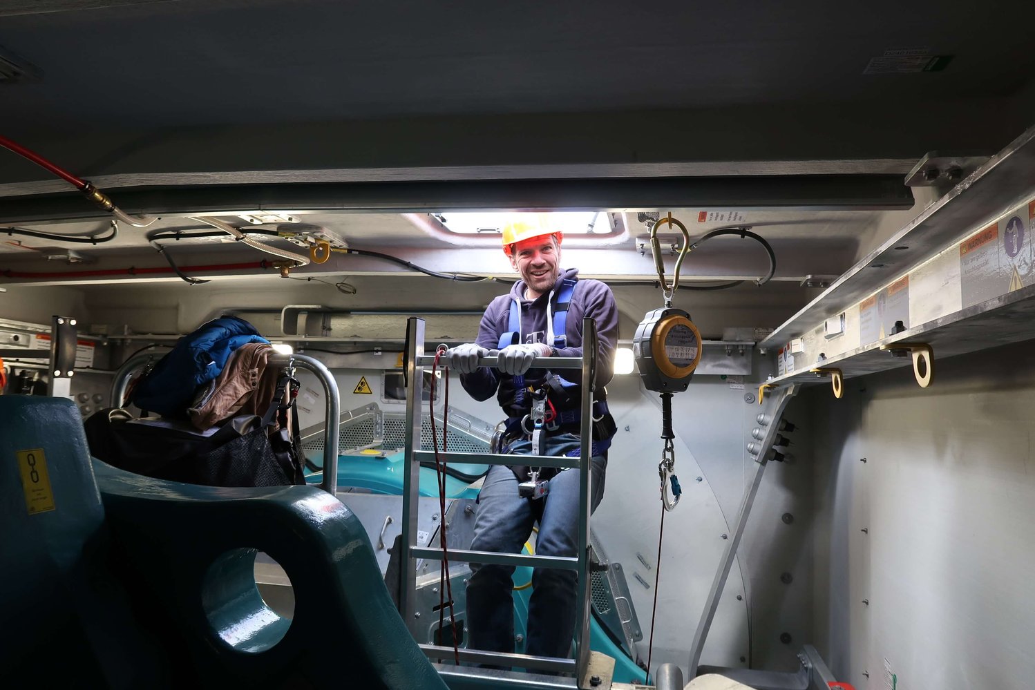 Eine Person mit Schutzhelm und Sicherheitsgurt steht in einer Industrieanlage auf einer Leiter. In der Nähe sind Ausrüstung und Taschen zu sehen.