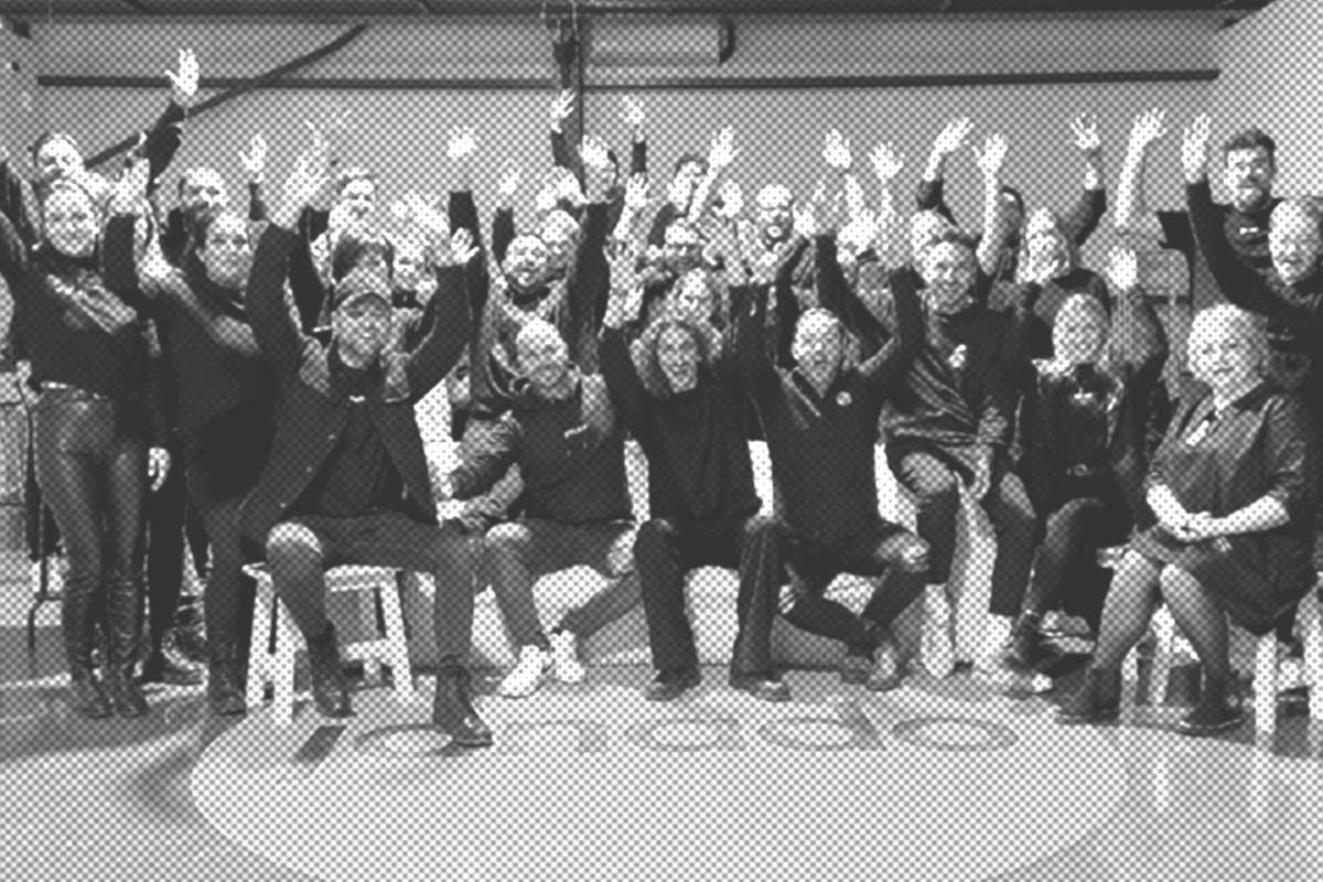 Eine Gruppe von Menschen sitzt und steht in einem Studio und streckt jeweils einen Arm in die Luft. Die meisten tragen dunkle Kleidung und scheinen für das Foto zu posieren.