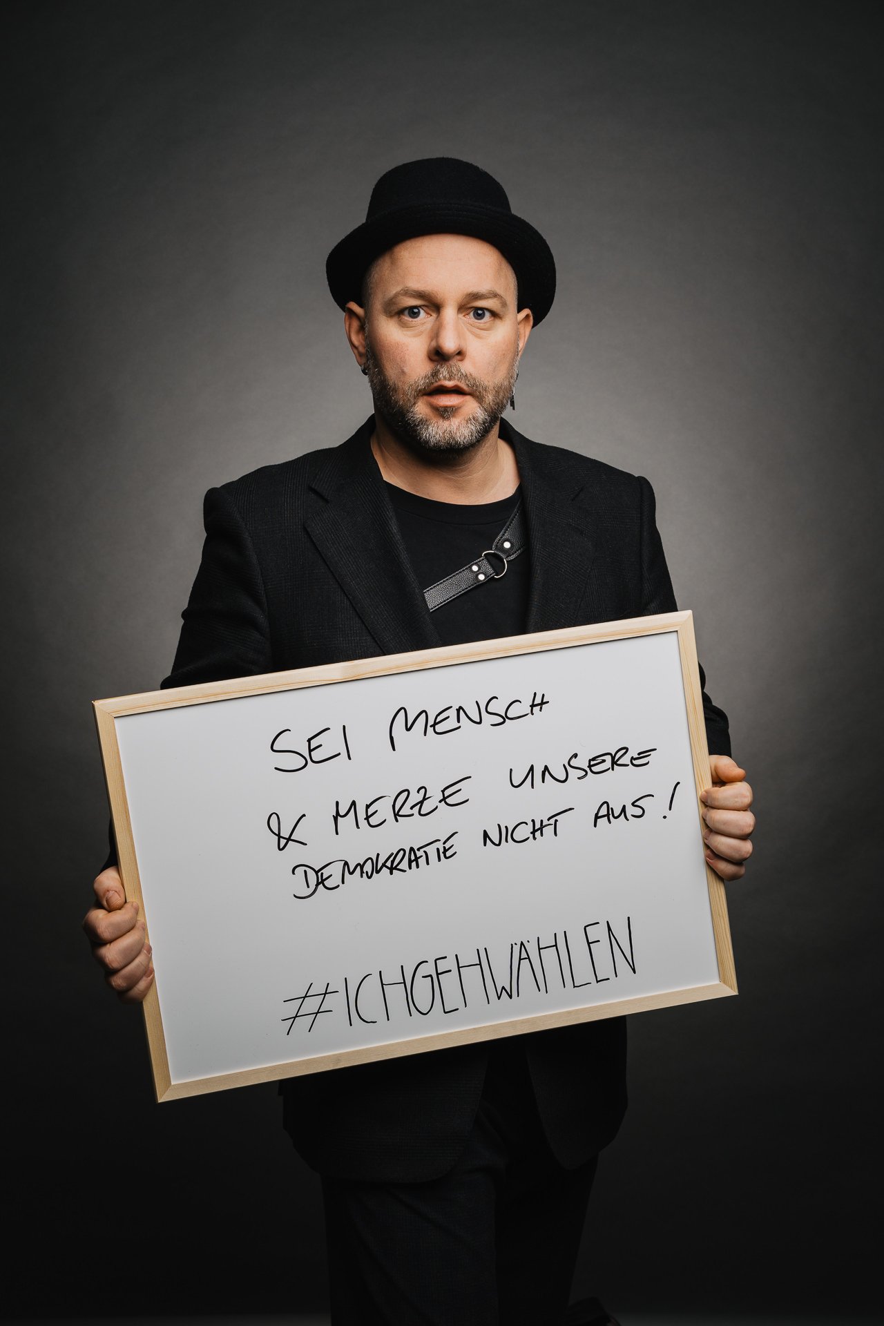 Person in schwarzem Outfit und Hut, die ein Schild mit einer Botschaft zur Demokratie hält, darunter der Hashtag #ichgehwählen.