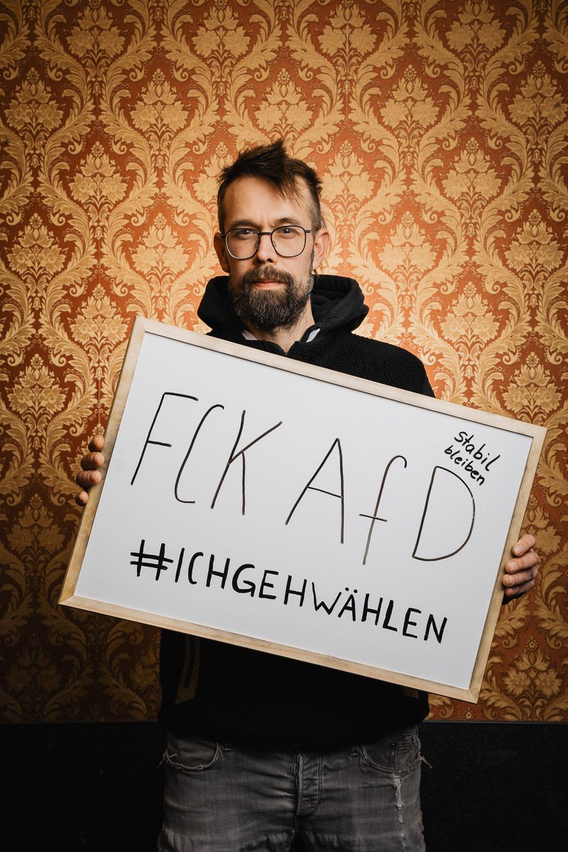 Person, die ein Schild mit einer Botschaft gegen eine politische Partei hält und vor einem verzierten Tapetenhintergrund steht.