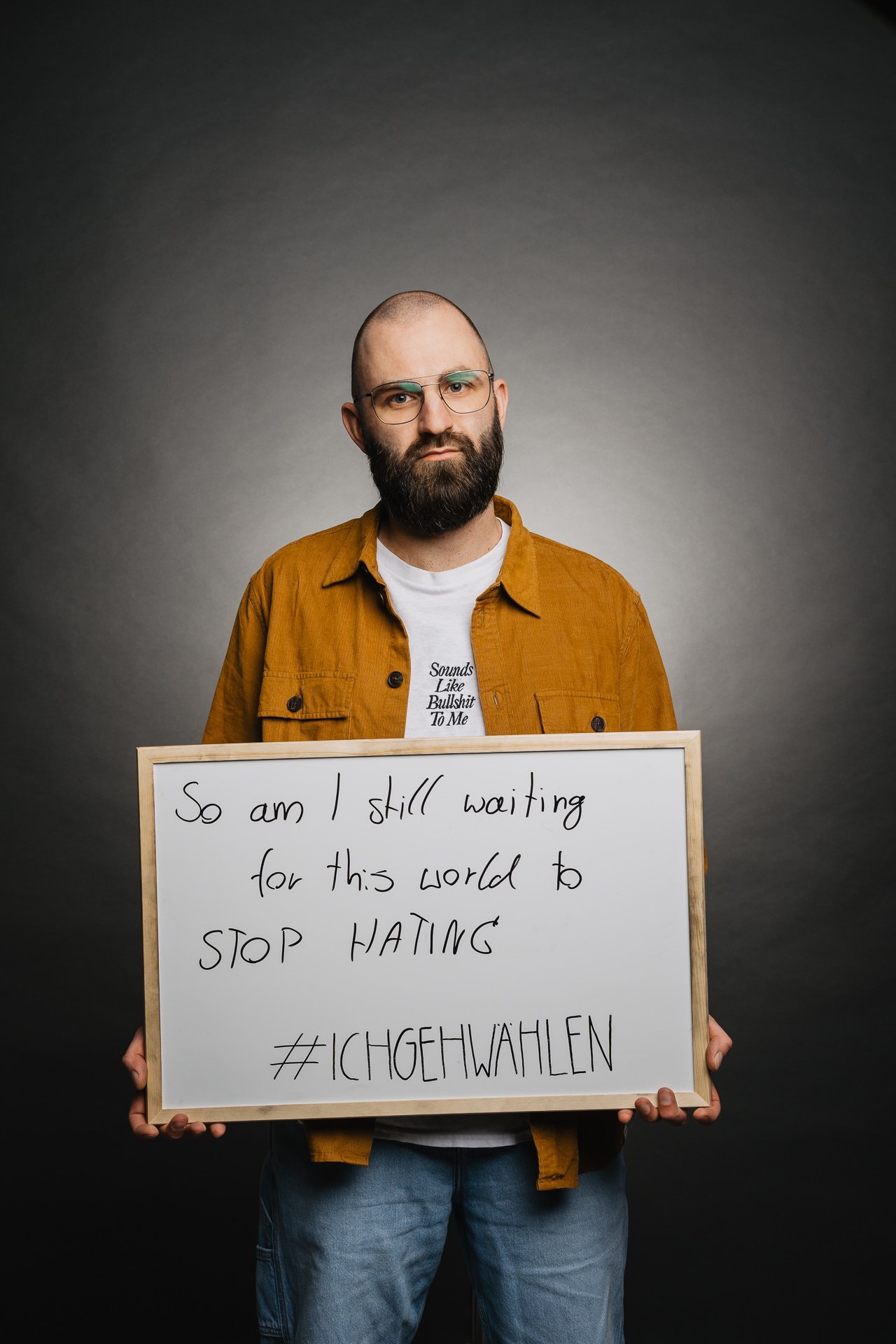 Eine Person mit Bart und Brille hält ein Schild mit der Aufschrift: „Also warte ich immer noch darauf, dass diese Welt aufhört zu hassen.“.