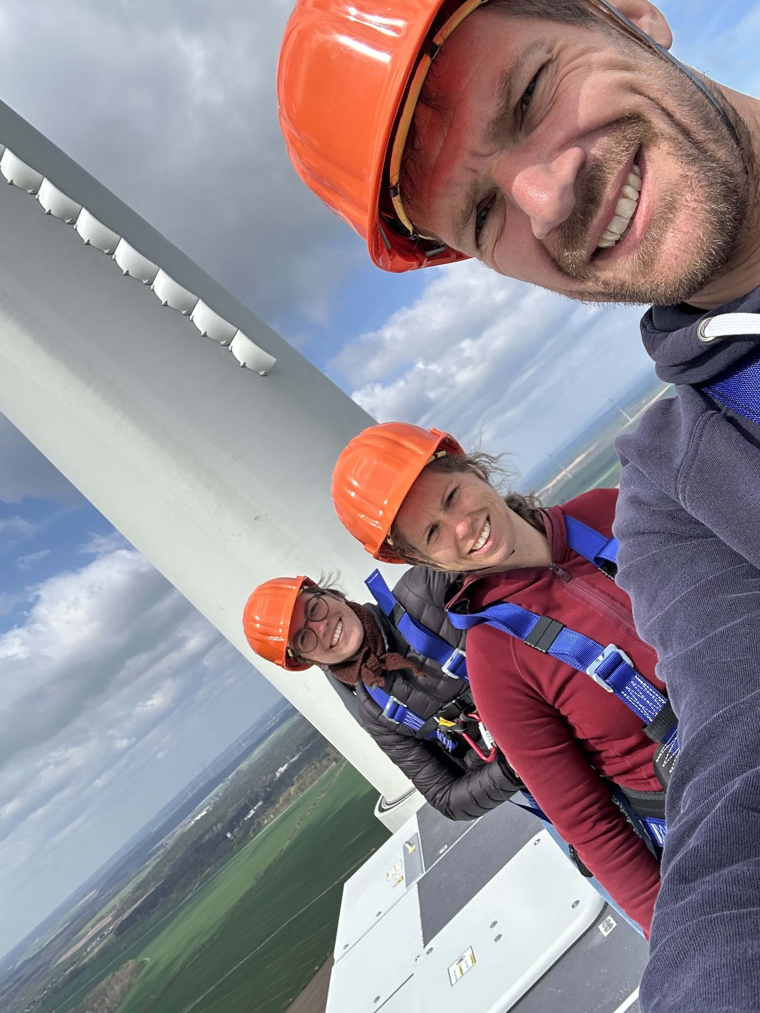 Eine Person in Schutzausrüstung und Helm steht in einem industriellen Metallaufzug oder -gehäuse und lächelt in die Kamera. Um sie herum sind Sicherheitsschilder und -ausrüstung sichtbar.