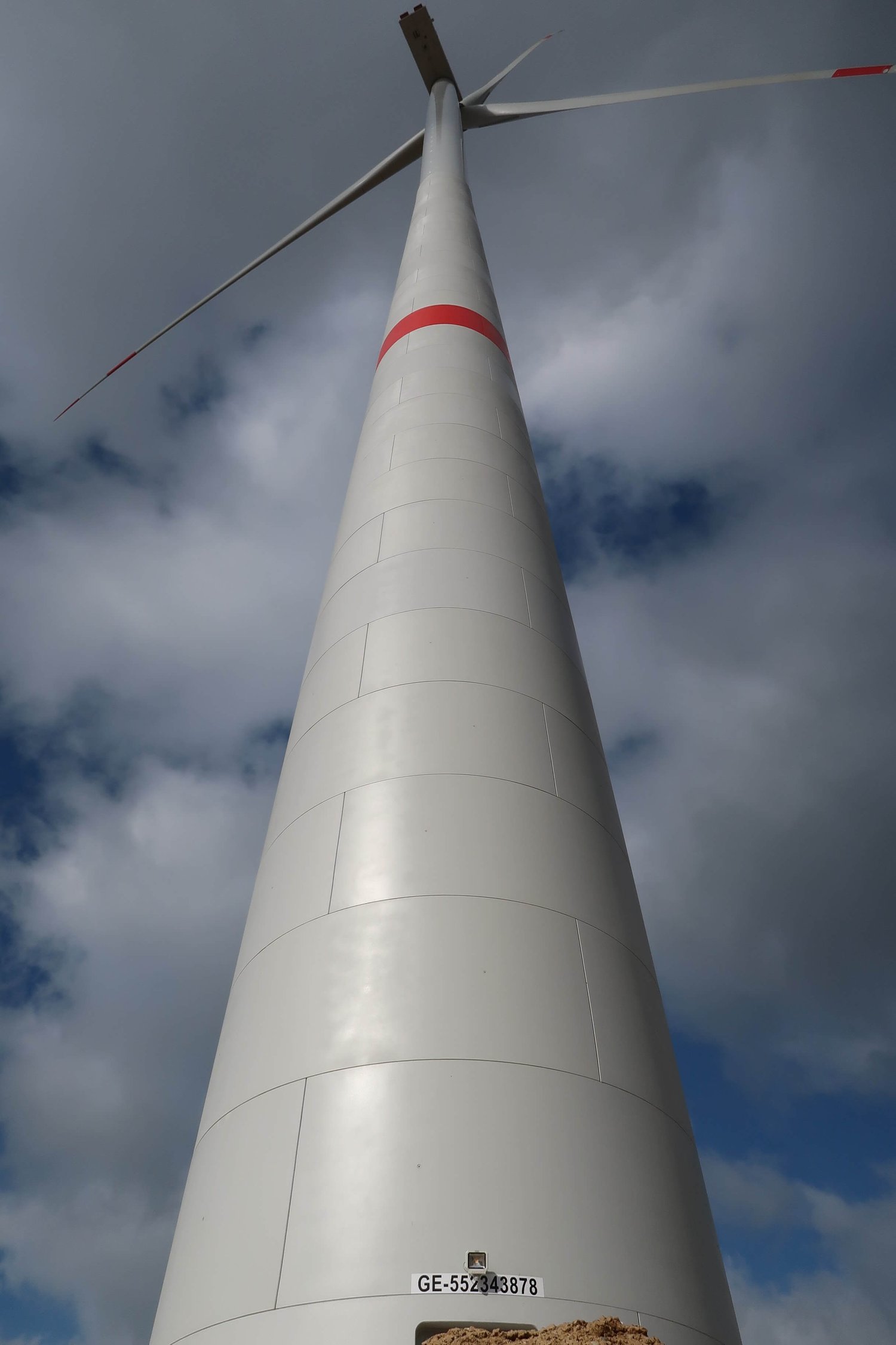 Eine Nahaufnahme einer Windkraftanlage von oben vor einem bewölkten Himmel. Man sieht ihren hohen weißen Turm mit einem roten Streifen und einer sichtbaren Identifikationsnummer in der Nähe der Basis.