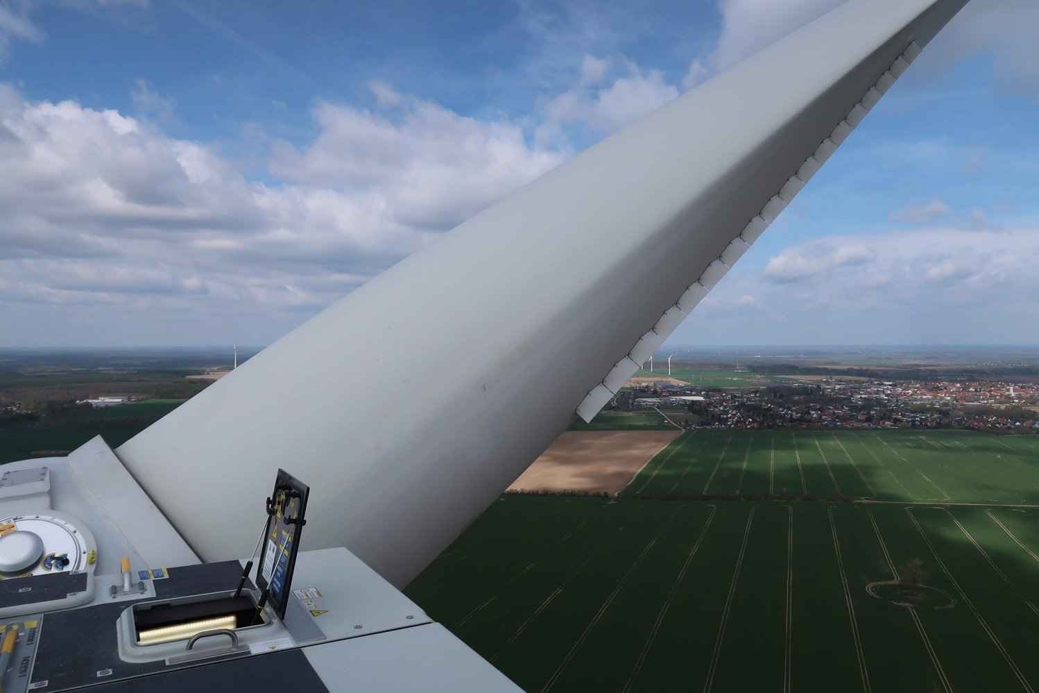 Ein Windturbinenblatt von der Spitze der Turbine aus gesehen, mit Blick auf grüne Felder und eine entfernte Stadt unter einem teilweise bewölkten Himmel.