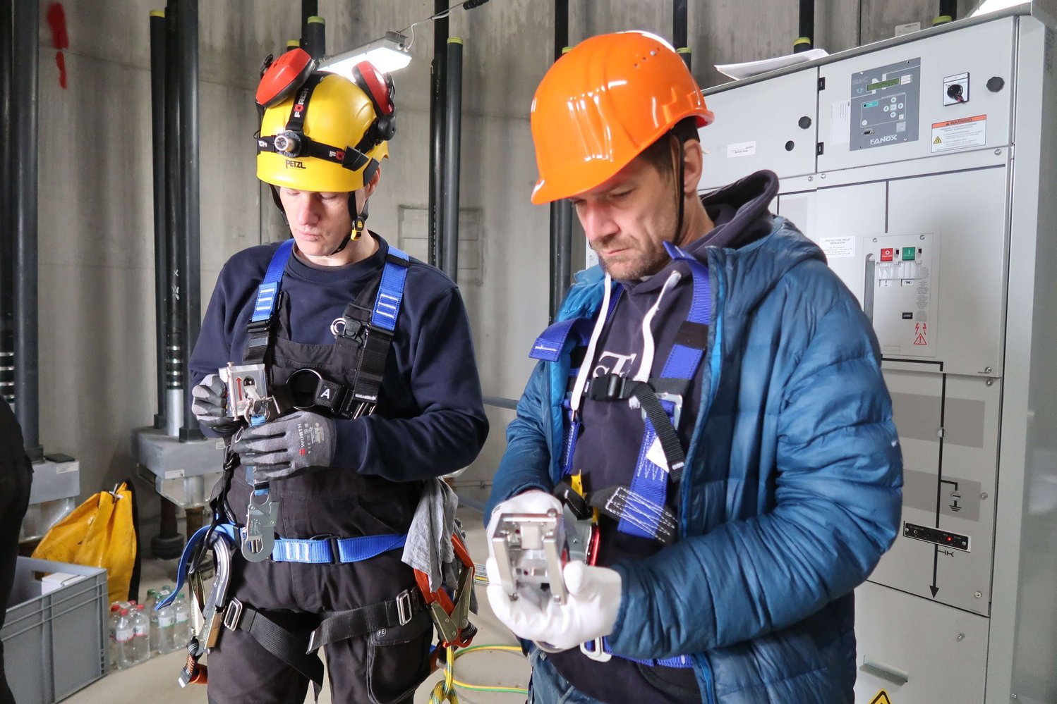 Zwei Arbeiter mit Helmen und Sicherheitsgurten untersuchen Handgeräte in einer Industrieanlage mit Schalttafeln im Hintergrund.