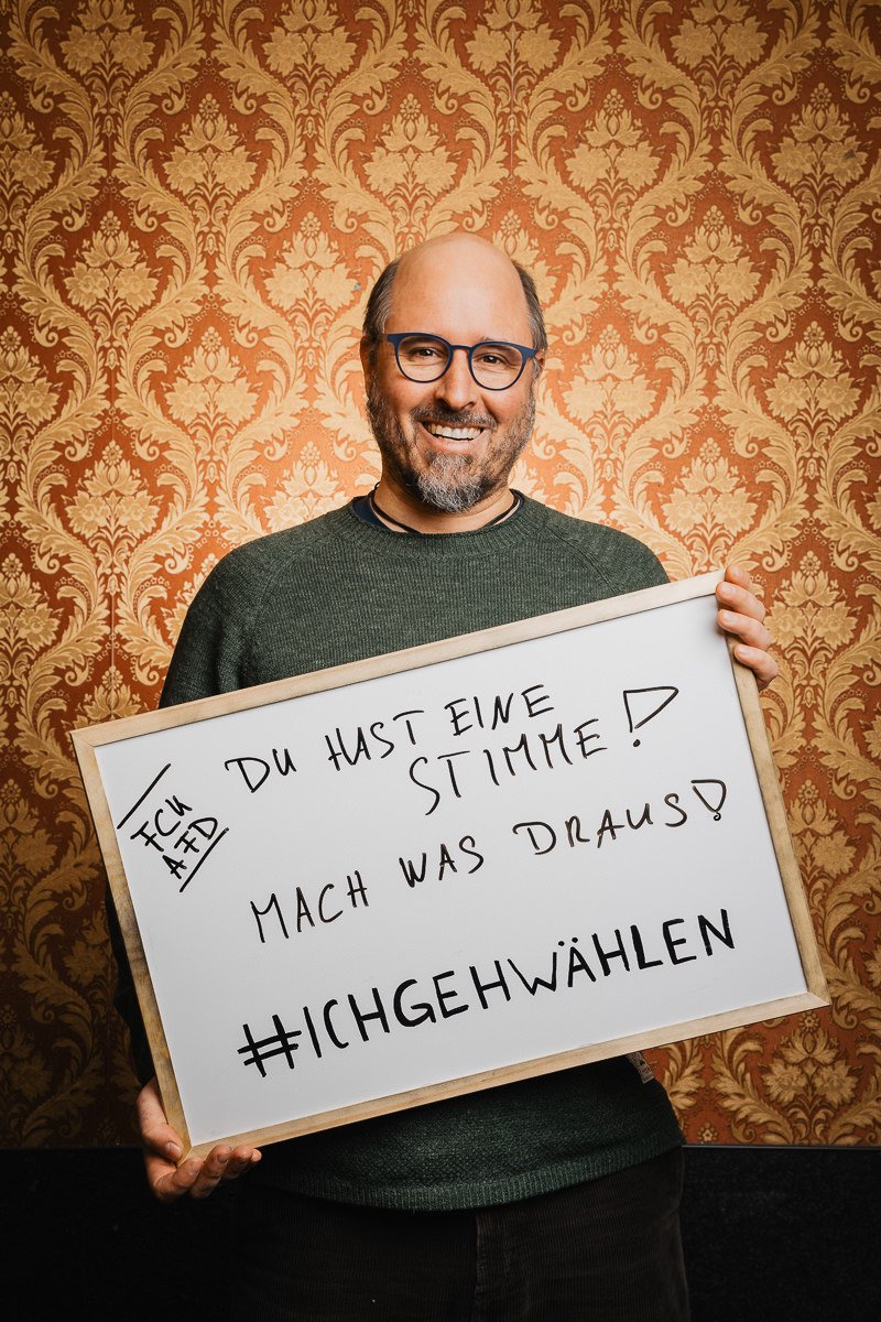 Ein Mann mit Brille hält ein Schild mit einer deutschen Botschaft, auf der zur Wahl aufgerufen wird, und steht vor einer gemusterten Tapete.