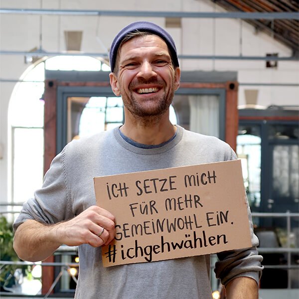 Ein Mann mit einer Mütze lächelt und hält ein Pappschild mit einem deutschen Text über Gemeinwohl und Wahlen. Im Hintergrund ein Innenraum mit Bogenfenstern.