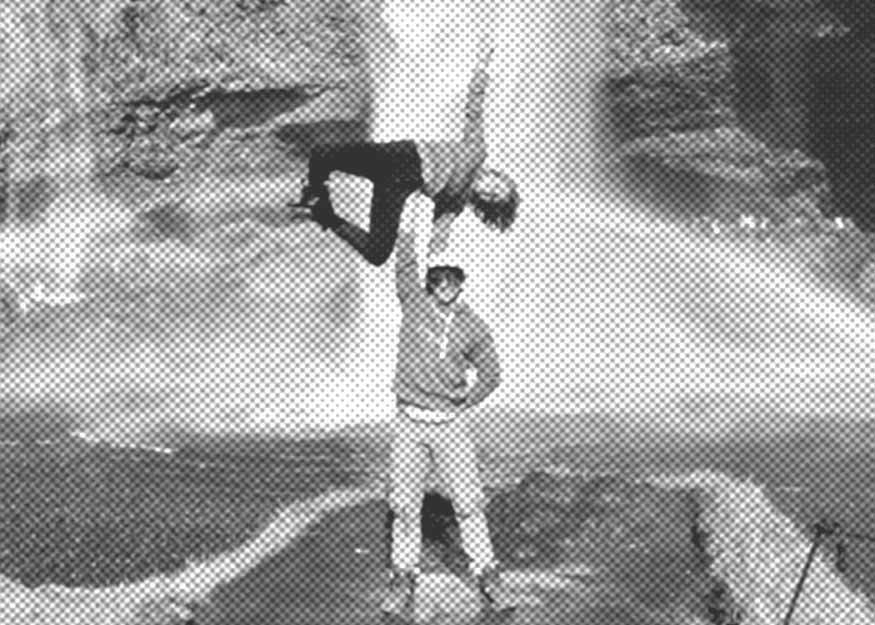 Ein Mann in Freizeitkleidung hebt vor einem Wasserfall eine Frau mit einer Hand über seinen Kopf.