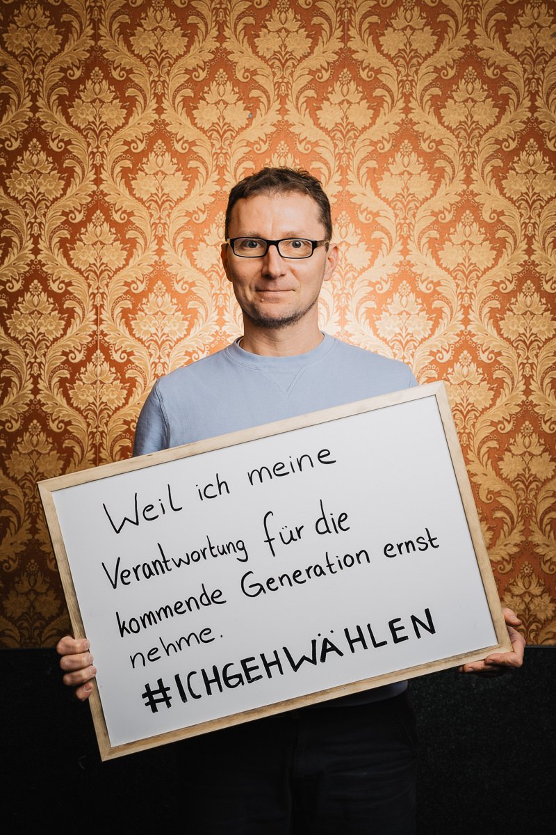 Eine Person hält vor einem gemusterten Tapetenhintergrund ein Schild mit einem handgeschriebenen Text auf Deutsch zur Wahlverantwortung.