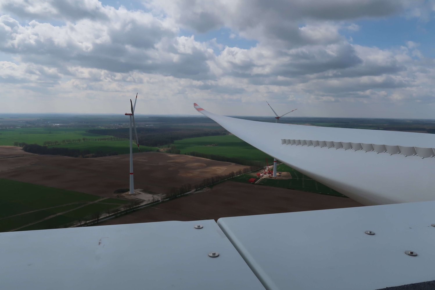 Blick von der Spitze einer Windkraftanlage mit Rotorblättern im Vordergrund, wobei unter einem teilweise bewölkten Himmel weitere Windkraftanlagen und Felder sichtbar sind.