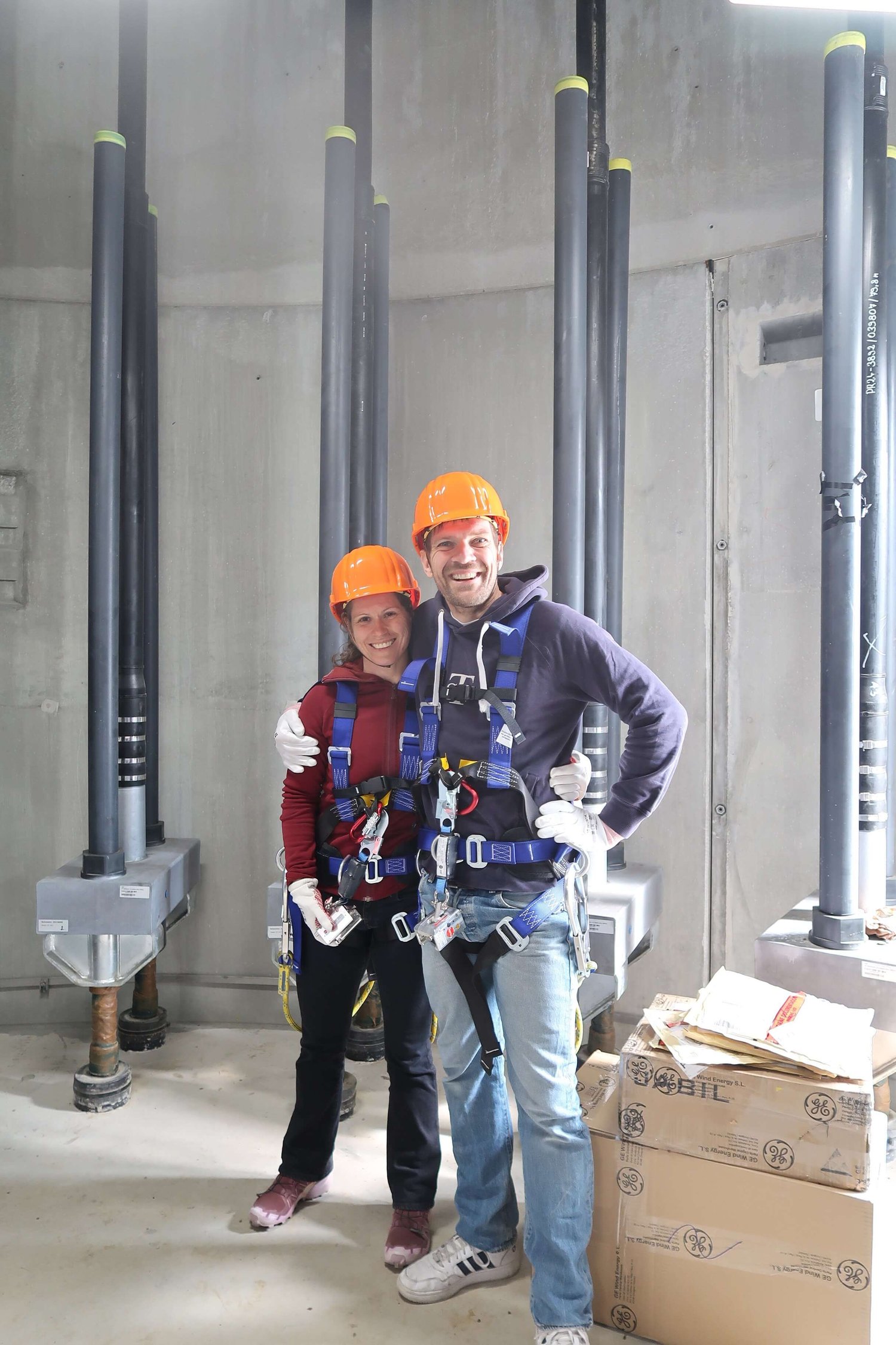 Zwei Personen mit Sicherheitsgurten und orangefarbenen Schutzhelmen stehen zusammen in einer Industrieanlage. In der Nähe stehen große Rohre und ein Karton.