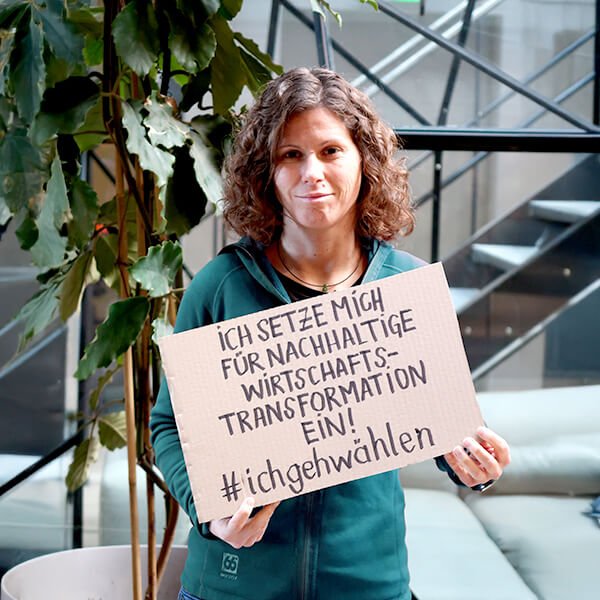 Eine Person hält ein Schild mit der Aufschrift „Ich stehe für eine nachhaltige wirtschaftliche Transformation!“ und „#ichgehwaehlen“ in der Hand und steht im Innenbereich neben einer Pflanze und einem Glasgeländer.