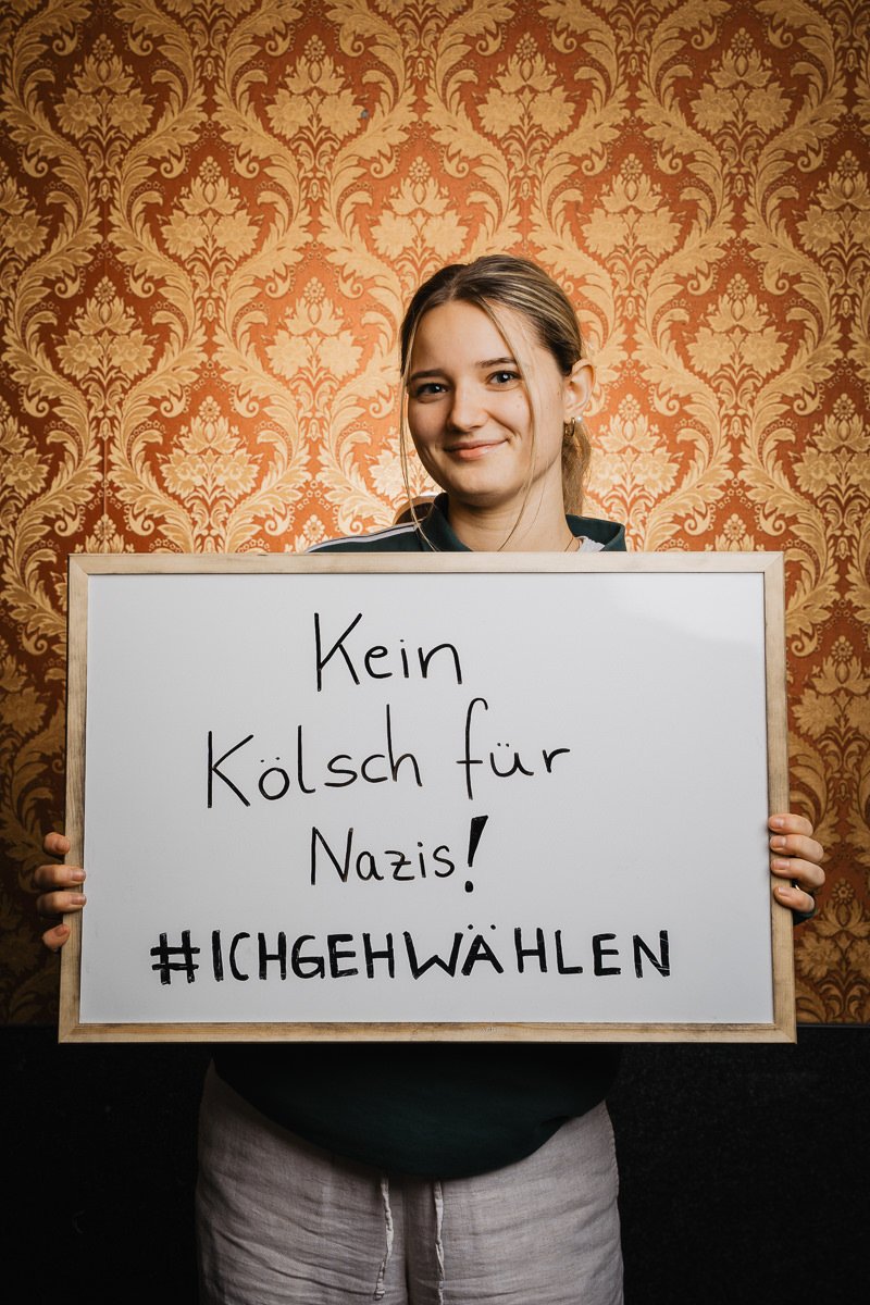 Person hält ein Schild mit der Aufschrift „Kein Kölsch für Nazis! #ICHGEHWÄHLEN“ vor einem gemusterten Tapetenhintergrund.