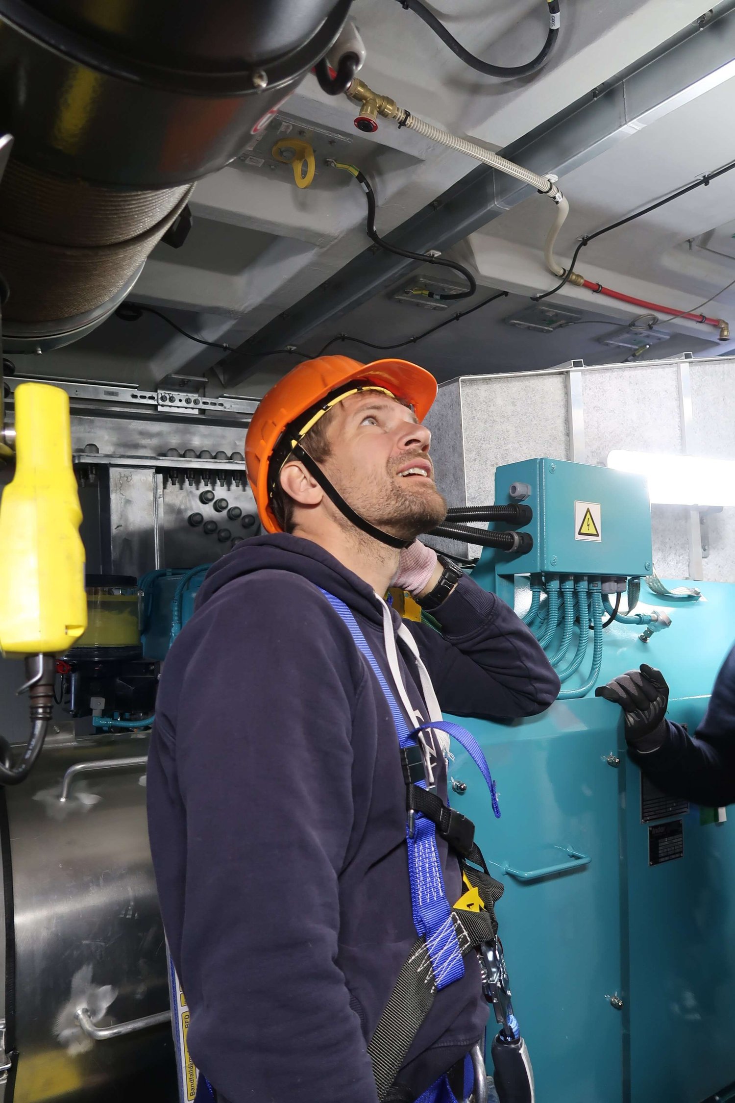 Eine Person mit Sicherheitsgurt, Handschuhen und orangefarbenem Schutzhelm steht im Innenbereich in der Nähe von Industrieanlagen und Sicherheitswarnschildern.
