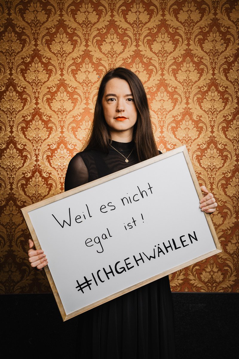 Person hält ein Schild mit der Aufschrift „Weil es nicht egal ist! #ICHGEHWÄHLEN“ vor einem gemusterten Tapetenhintergrund.