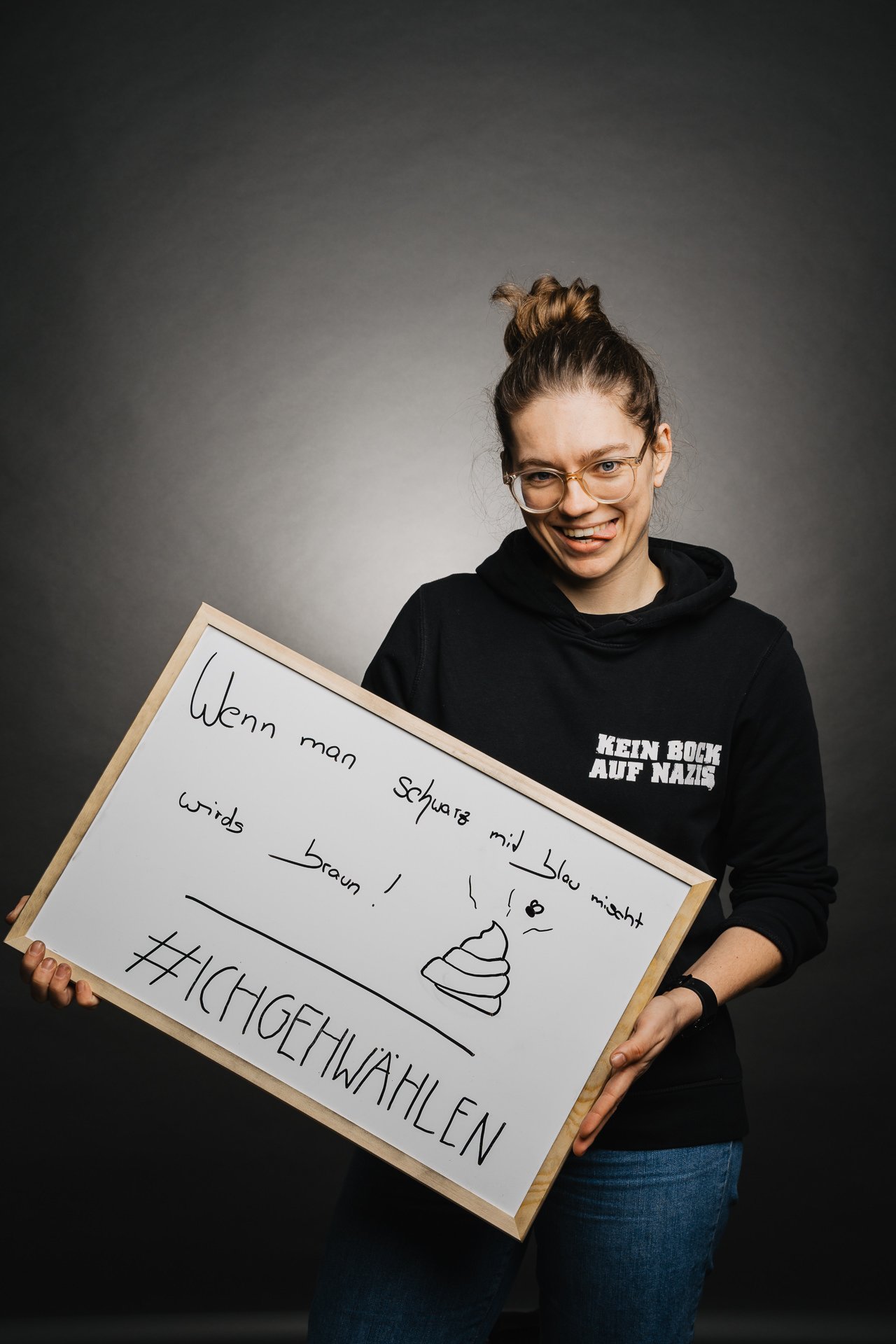 Person hält ein Schild mit deutschem Text und der Zeichnung eines Kackhaufen-Emojis. Sie trägt eine Brille und einen schwarzen Kapuzenpullover mit Text.
